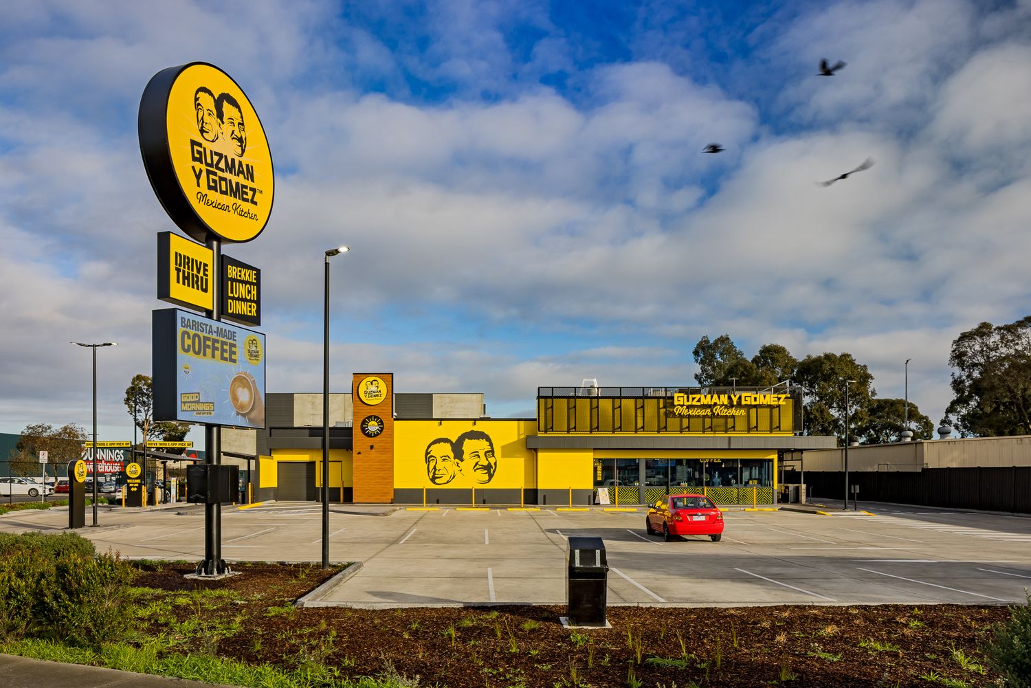 Guzman y Gomez restaurant with tall roadside sign, drive-through lane and a red car parked in the lot on a sunny day.