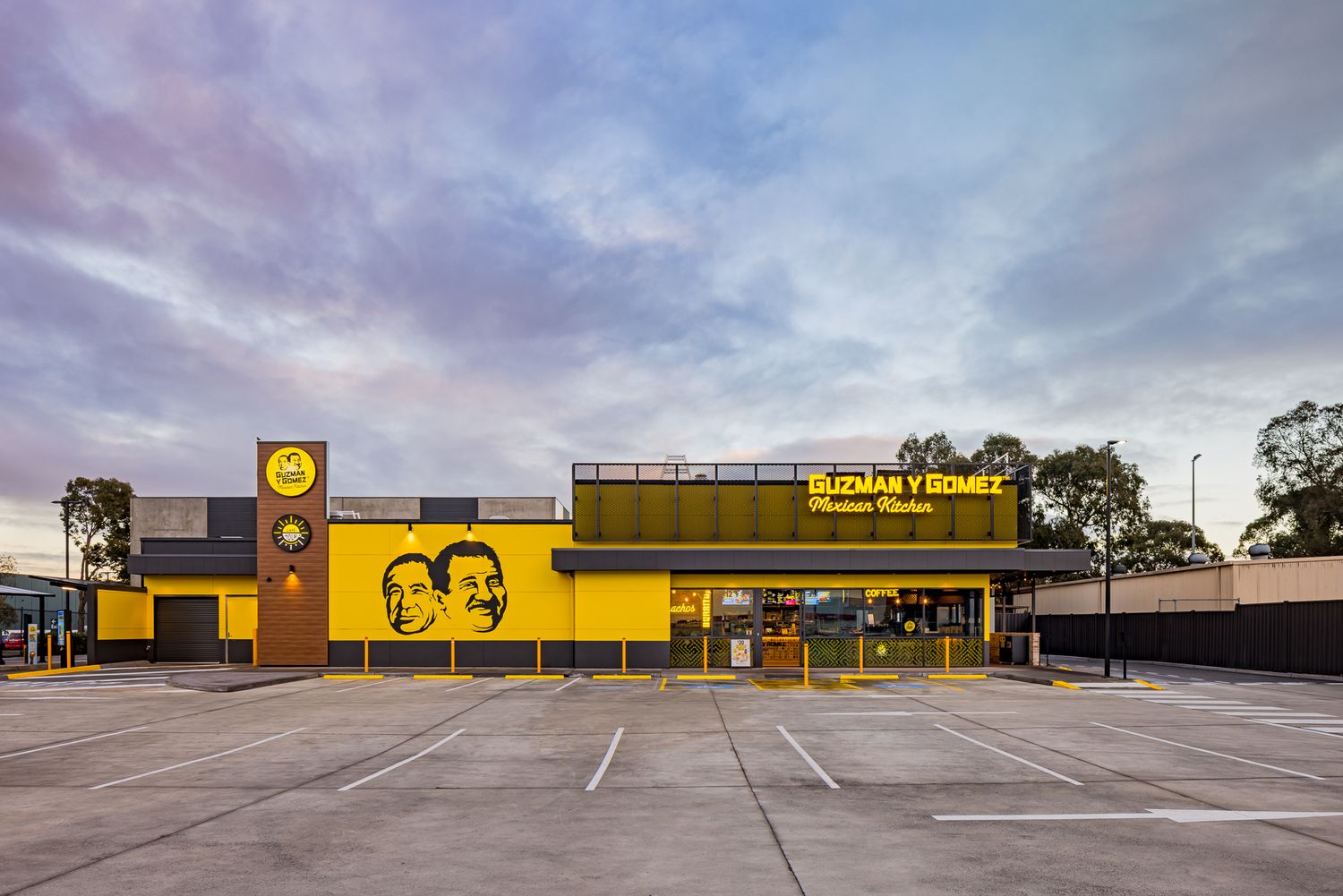 Guzman y Gomez Mexican Kitchen storefront with a yellow facade, illuminated sign and an empty parking lot under a pastel evening sky.