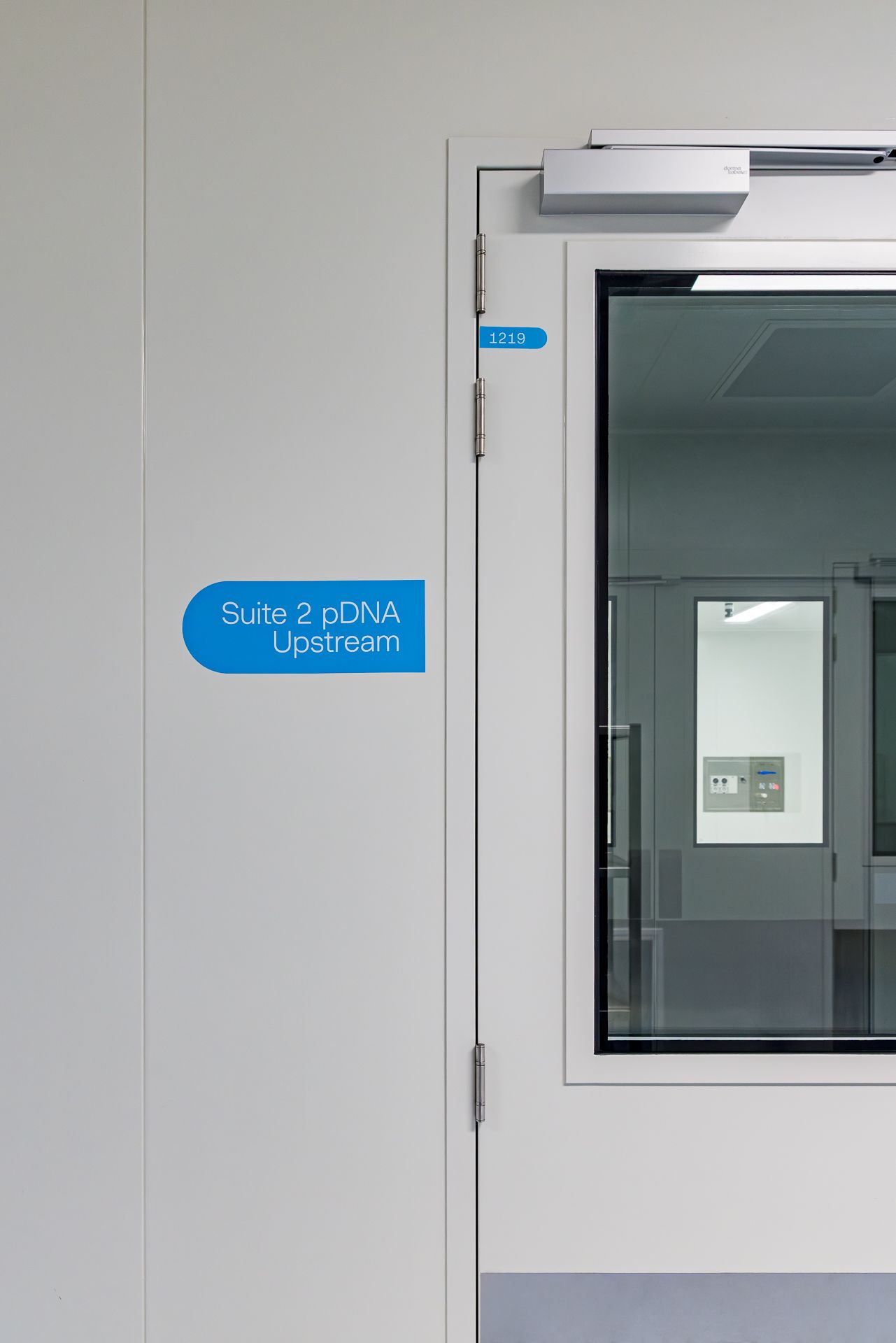 Cleanroom door and blue sign reading Suite 2 pDNA Upstream, with a glass window revealing a sterile processing room.