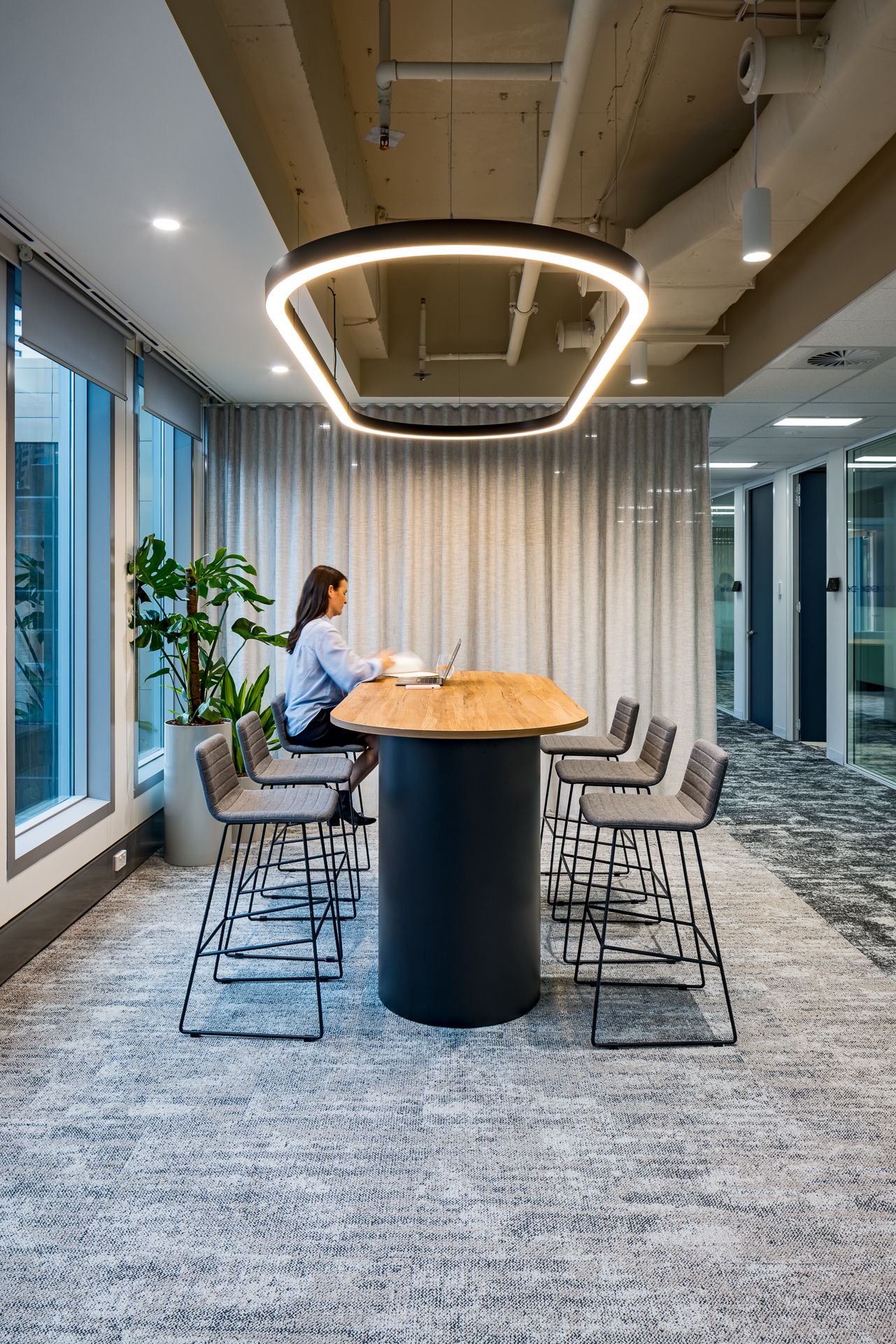 Woman working on a laptop at a high timber table with stools in a modern office, lit by a large circular pendant light.