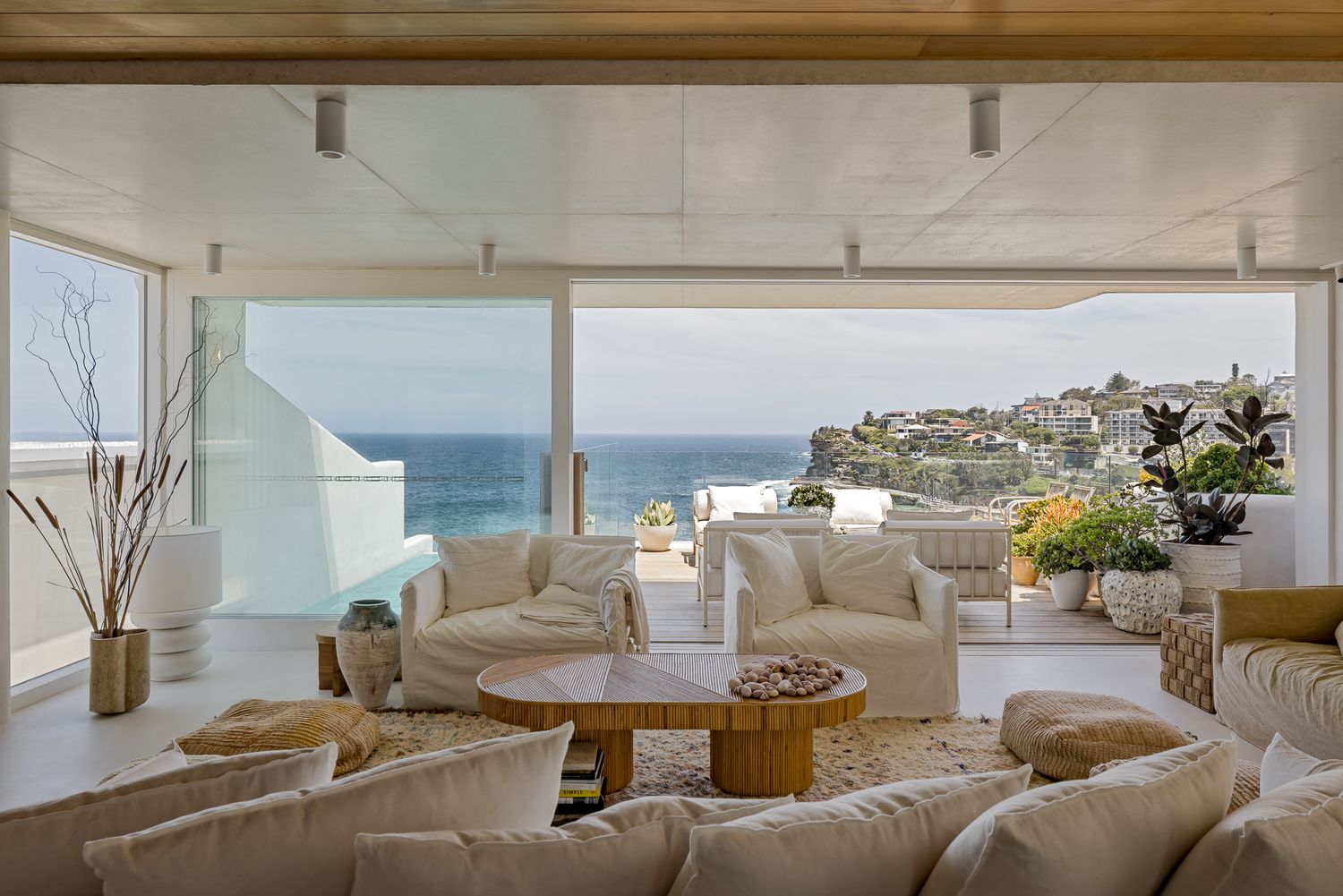 Open-plan coastal living room with cream sofas, a round timber coffee table and floor-to-ceiling windows overlooking the ocean.