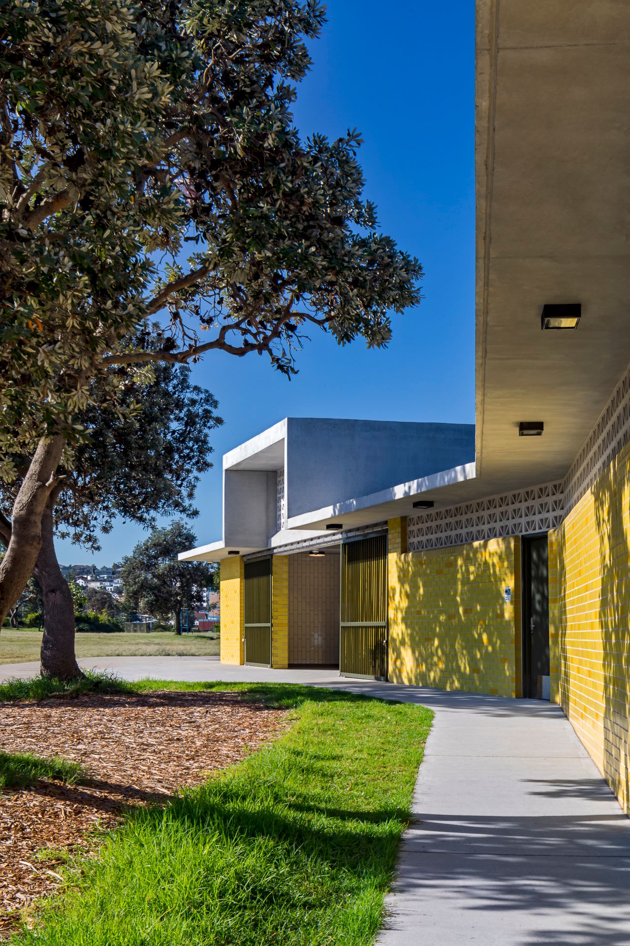 Concrete path leads along a yellow brick amenities building with breeze block detail, shaded by large trees and edged with green lawn.