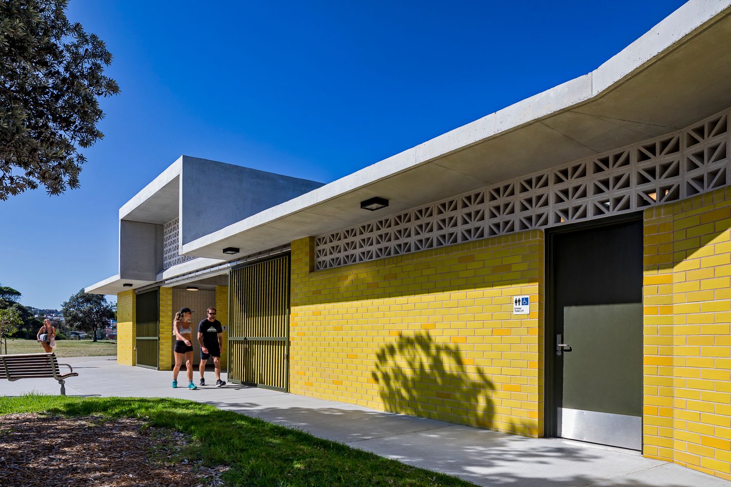 Two people stand at the entrance of a yellow brick amenities building featuring a patterned concrete breeze block screen along the roofline.