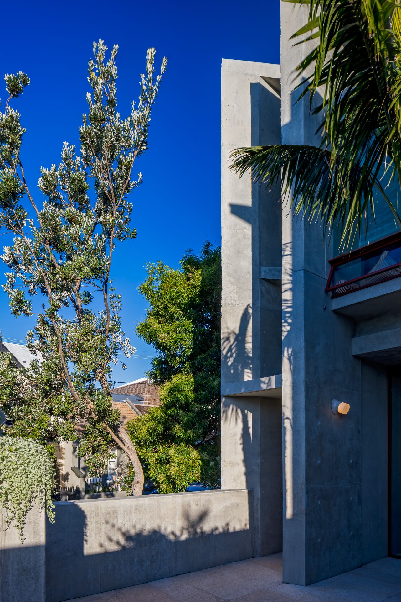 Sunlit concrete building with stepped walls beside a garden wall, a flowering tree, and palm fronds against a blue sky.