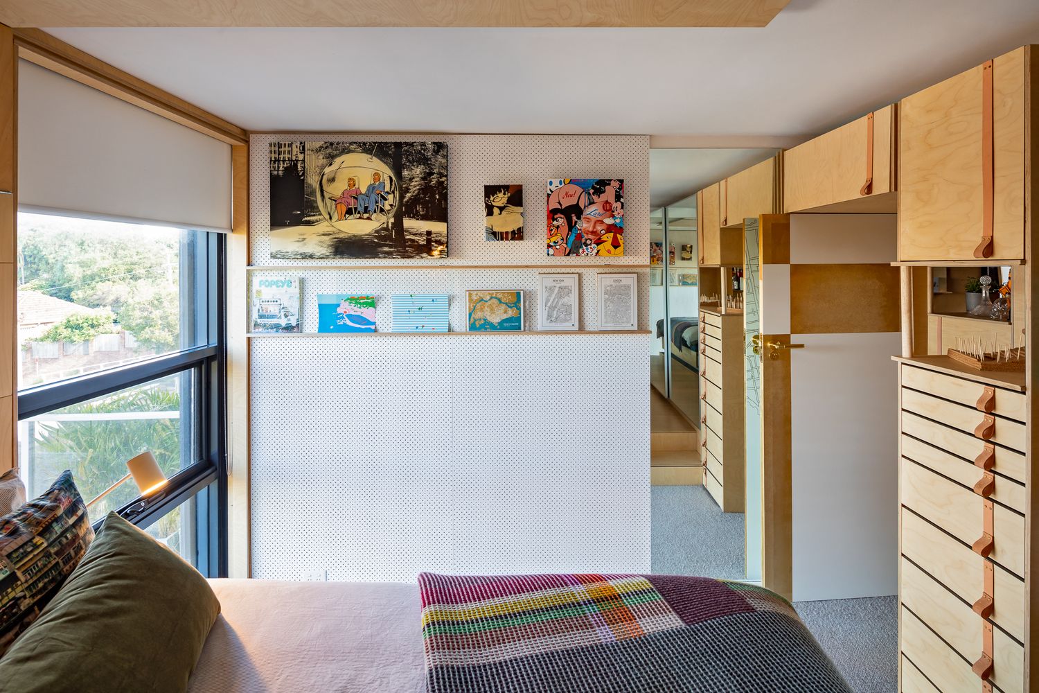 Plywood bedroom nook with a window, framed artwork shelf, wool blanket, and built-in drawers and cabinets.