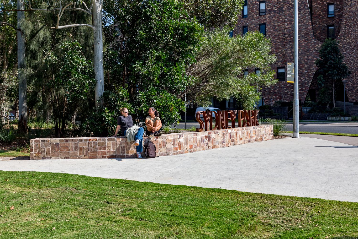 Two people sit on a sandstone wall behind rusted steel Sydney Park lettering beside a lawn and brick building.