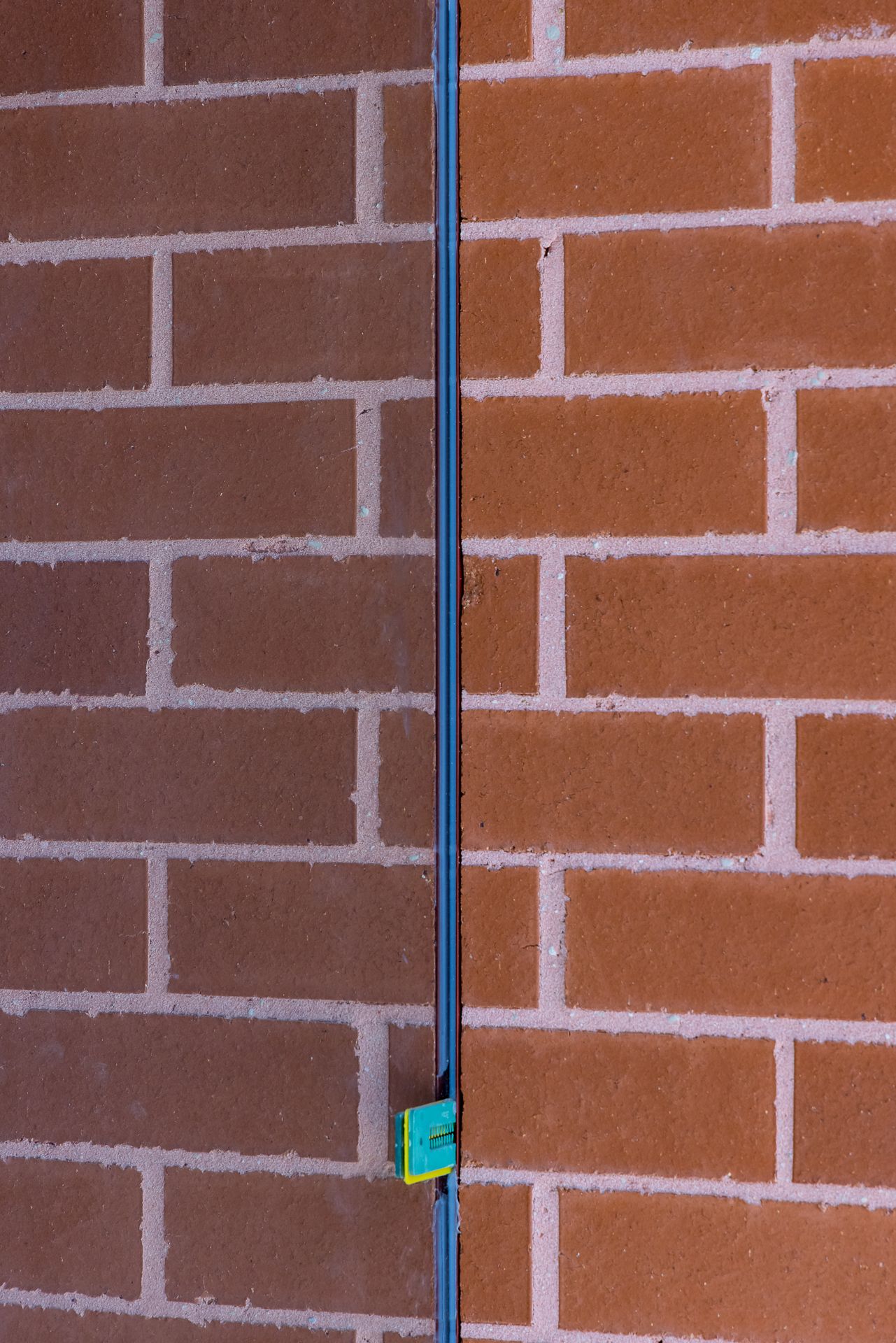 Detail of a red brick wall with light mortar joints split by a vertical blue metal strip and a small green and yellow bracket.