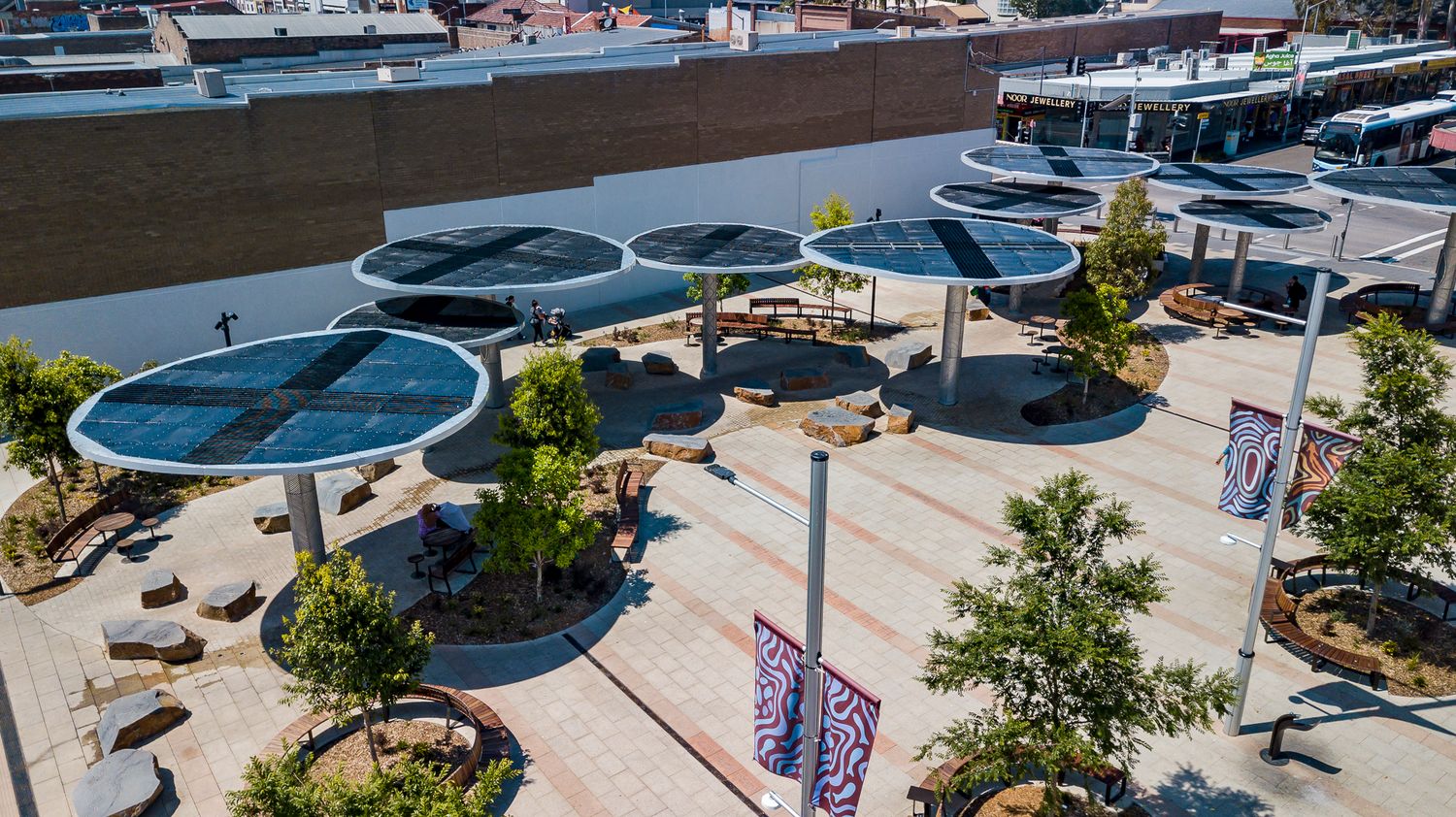 Elevated view of solar-panel shade structures, banners and young trees in a landscaped pedestrian plaza.