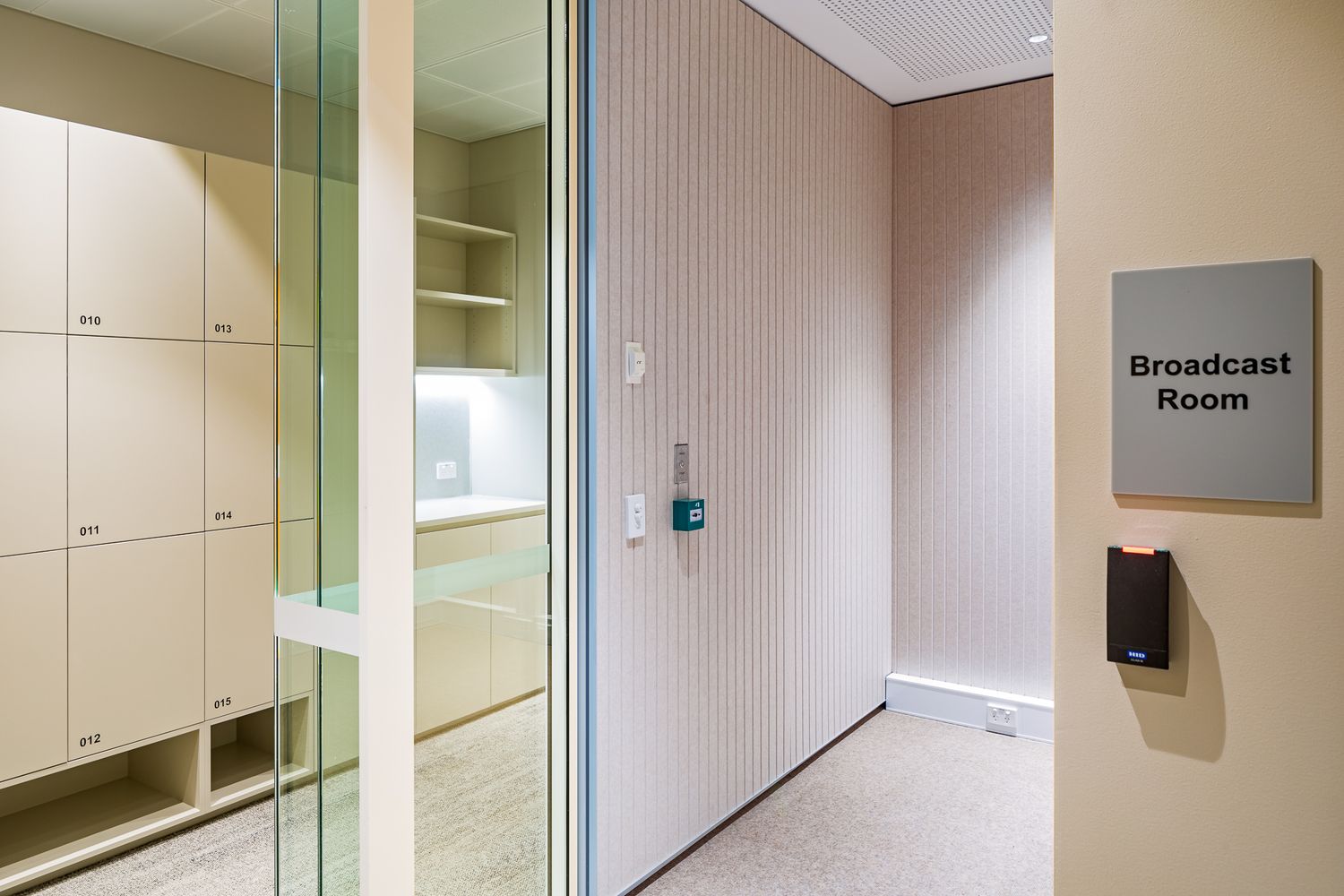 Office corridor with glass partitions, white storage lockers, slatted wood wall panels and a Broadcast Room door sign.
