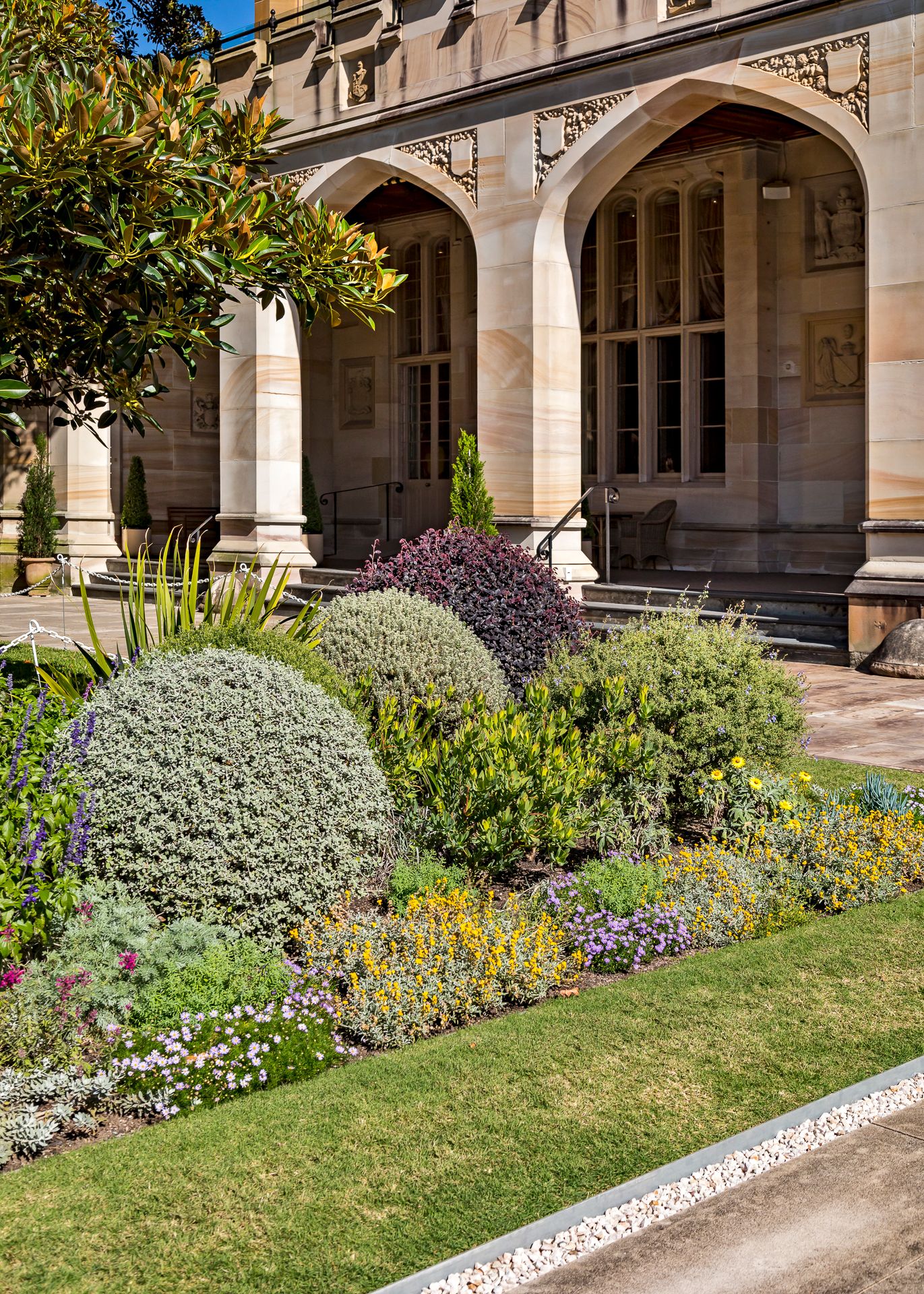 Colorful flower beds and rounded shrubs in front of a sandstone building with arched colonnades on a sunny day.