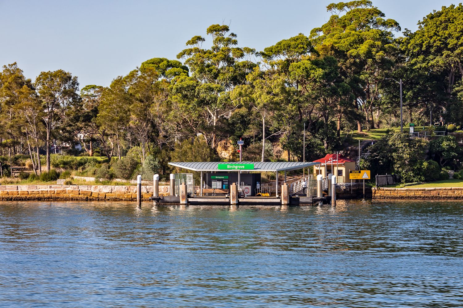 Small ferry wharf with a green sign and covered shelter sits at the water's edge beneath tall eucalyptus trees.