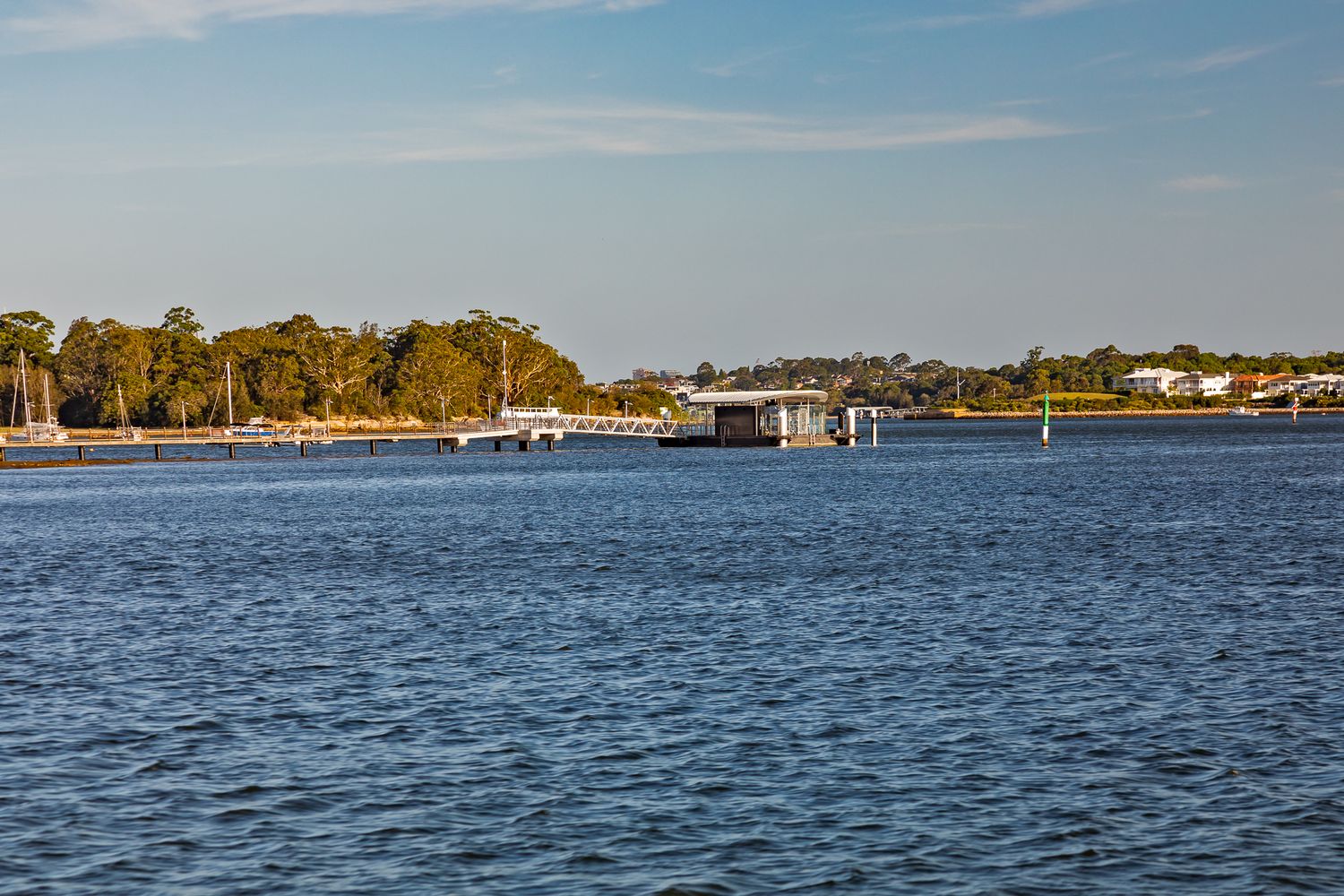 Calm blue harbour waters stretch toward a distant ferry wharf and tree-lined shoreline under a clear afternoon sky.