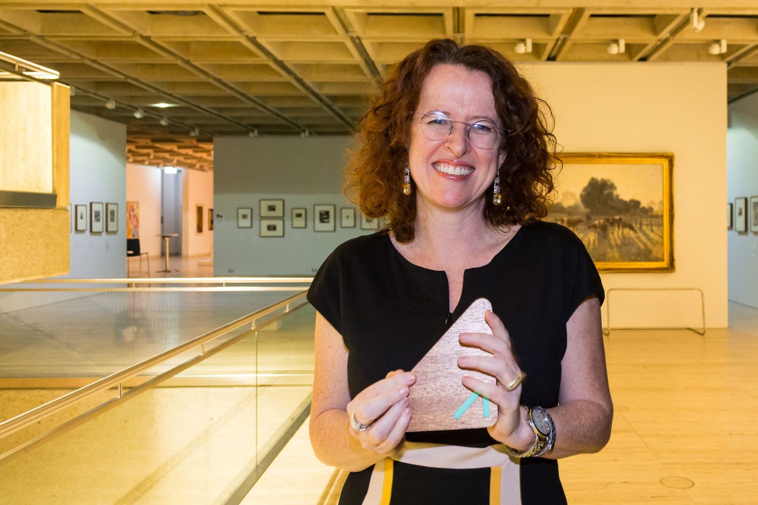 Smiling woman with curly hair and glasses holds a small pink object inside a bright art gallery with framed paintings.