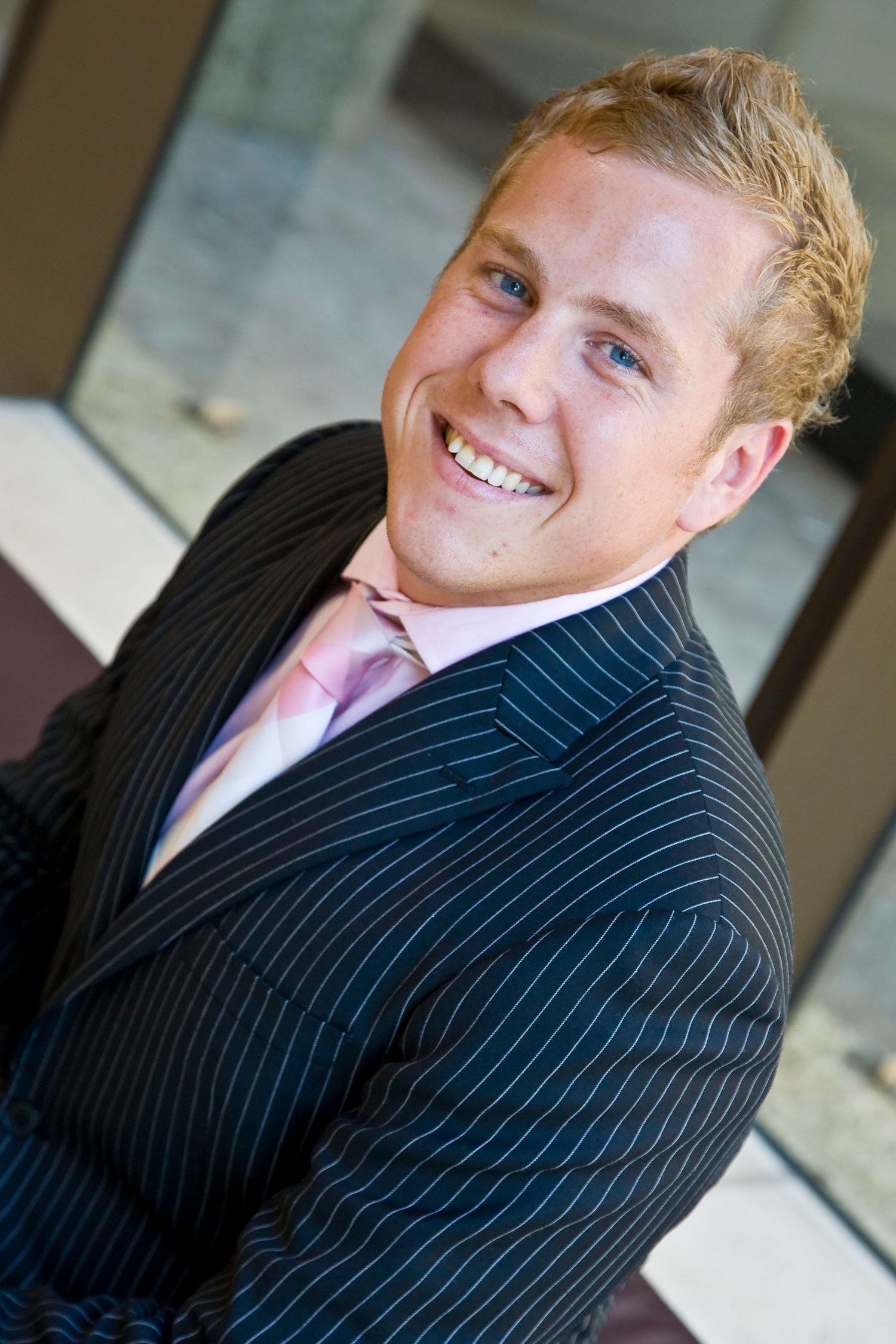 A smiling young man with blond hair wears a pinstripe suit and pink tie in a professional portrait.