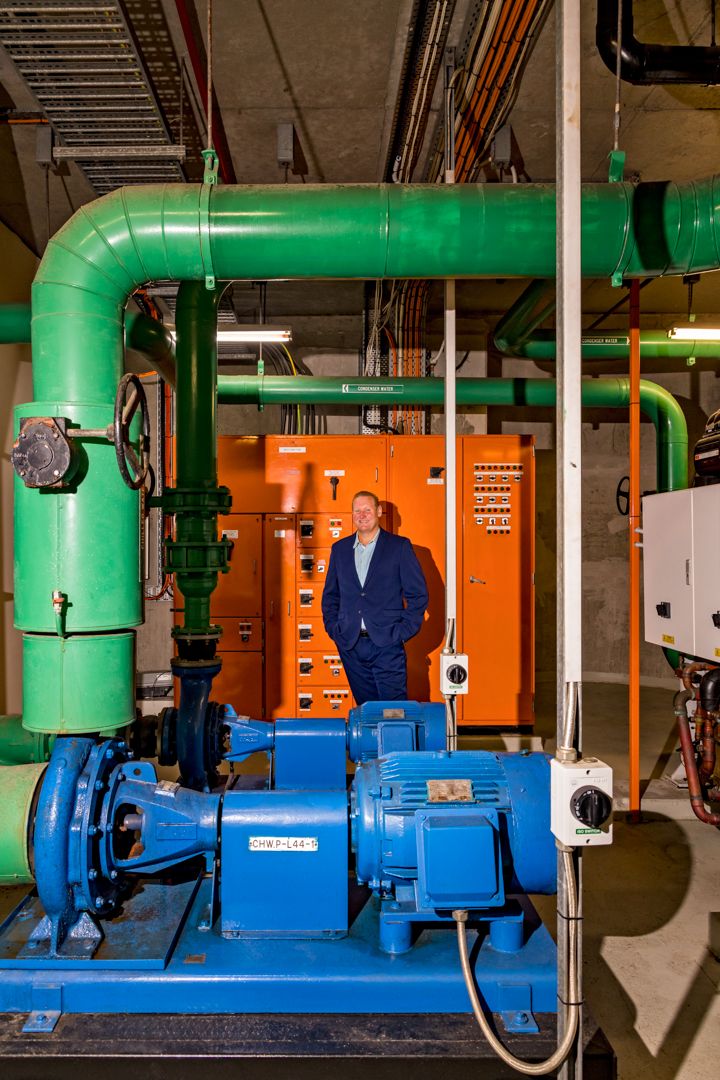 Man in a navy suit stands in a plant room among large green pipes, blue water pumps and an orange electrical control cabinet.
