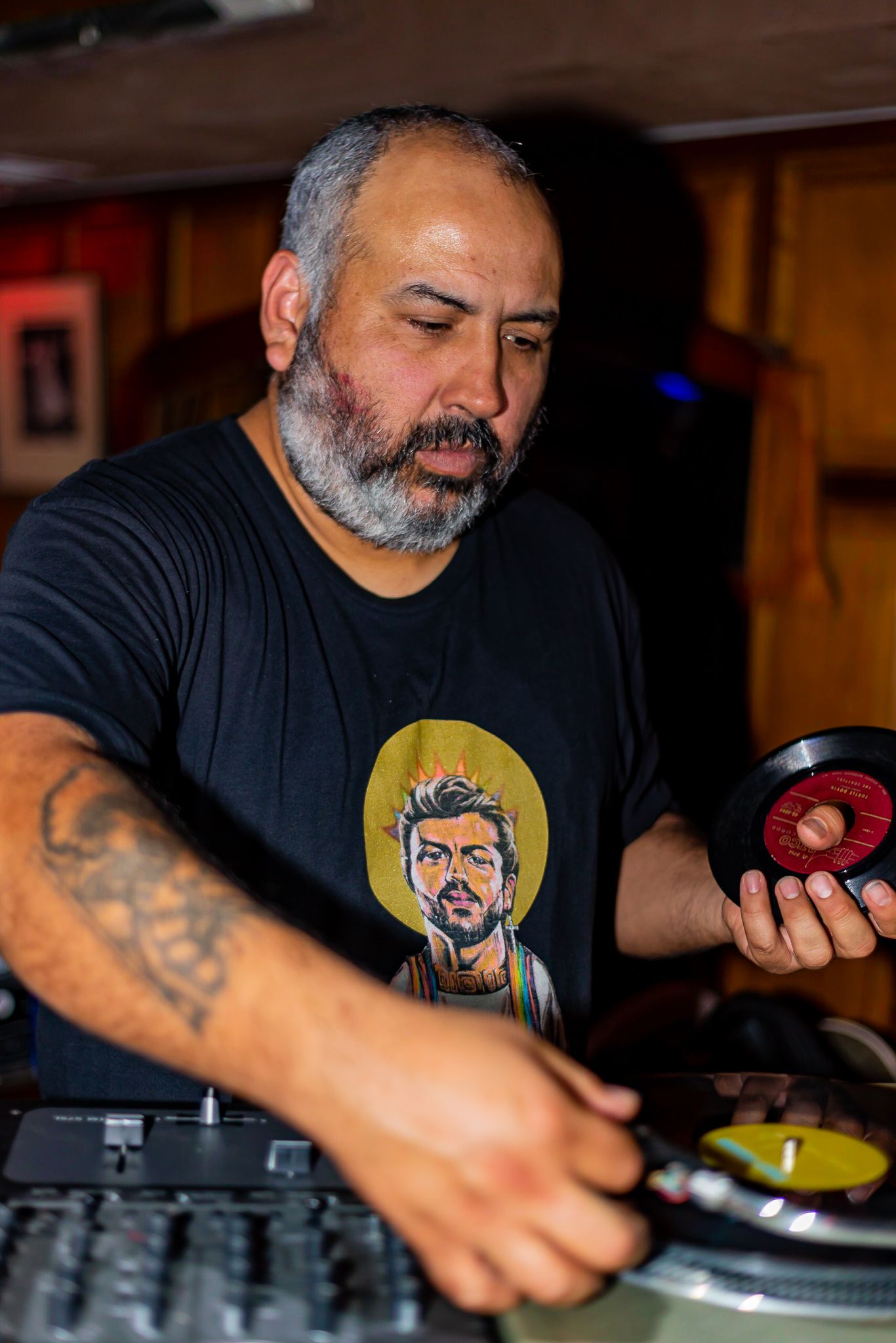 Bearded DJ in a black graphic t-shirt holds a vinyl record while mixing on turntables in a dimly lit, wood-paneled venue.