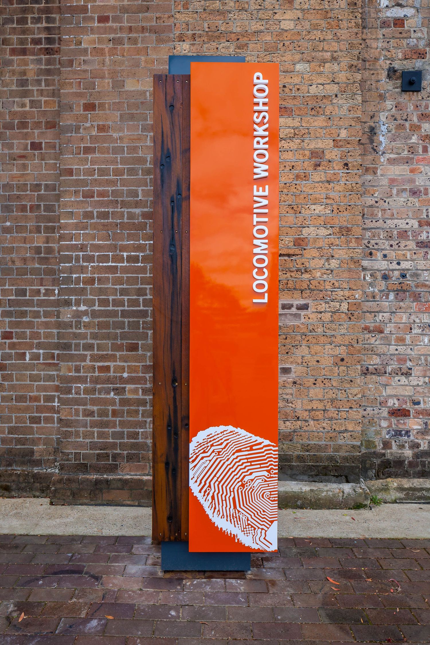 A tall orange Locomotive Workshop sign with a large fingerprint graphic stands against an old red brick wall.