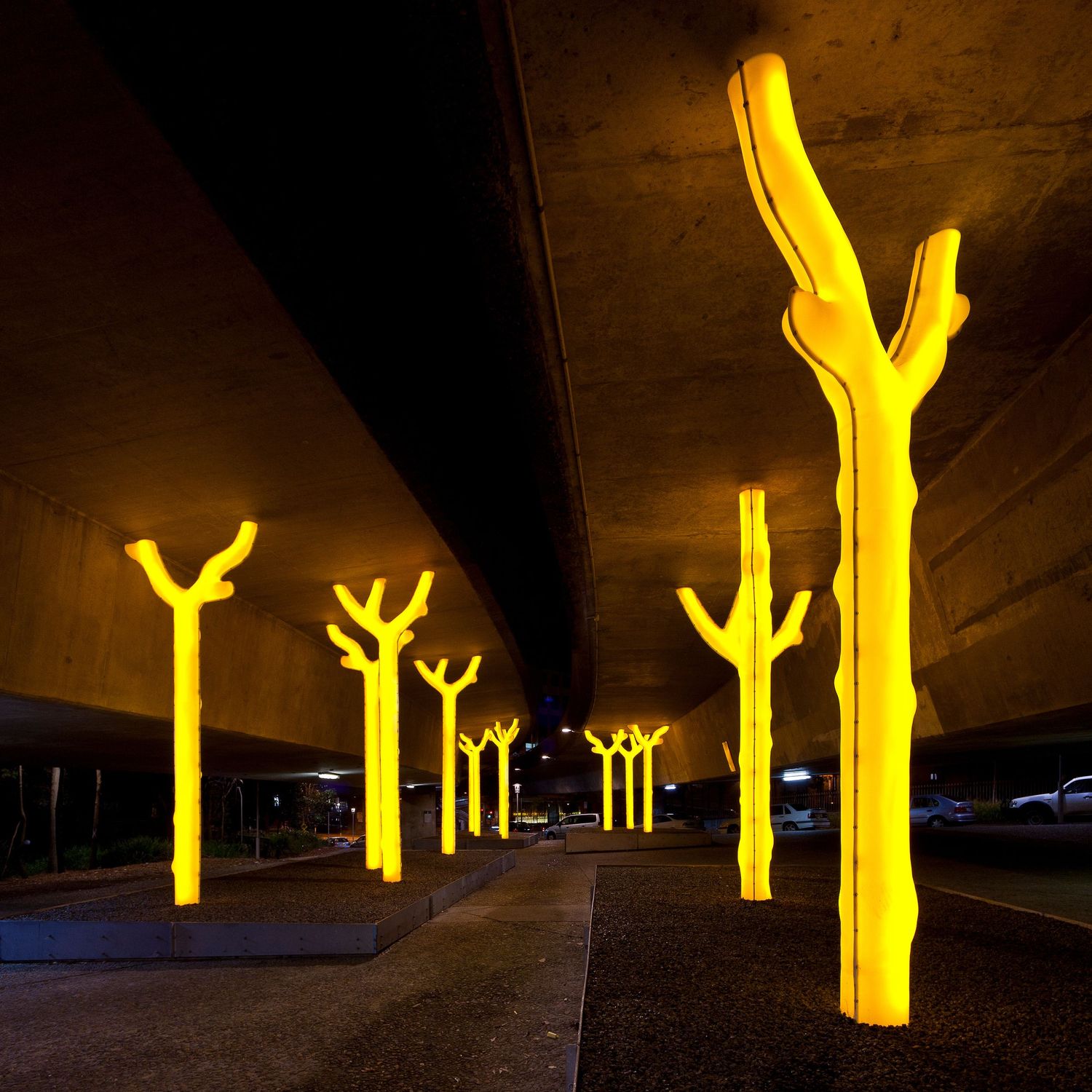 Glowing yellow tree-shaped light sculptures illuminating the space beneath a concrete overpass at night.