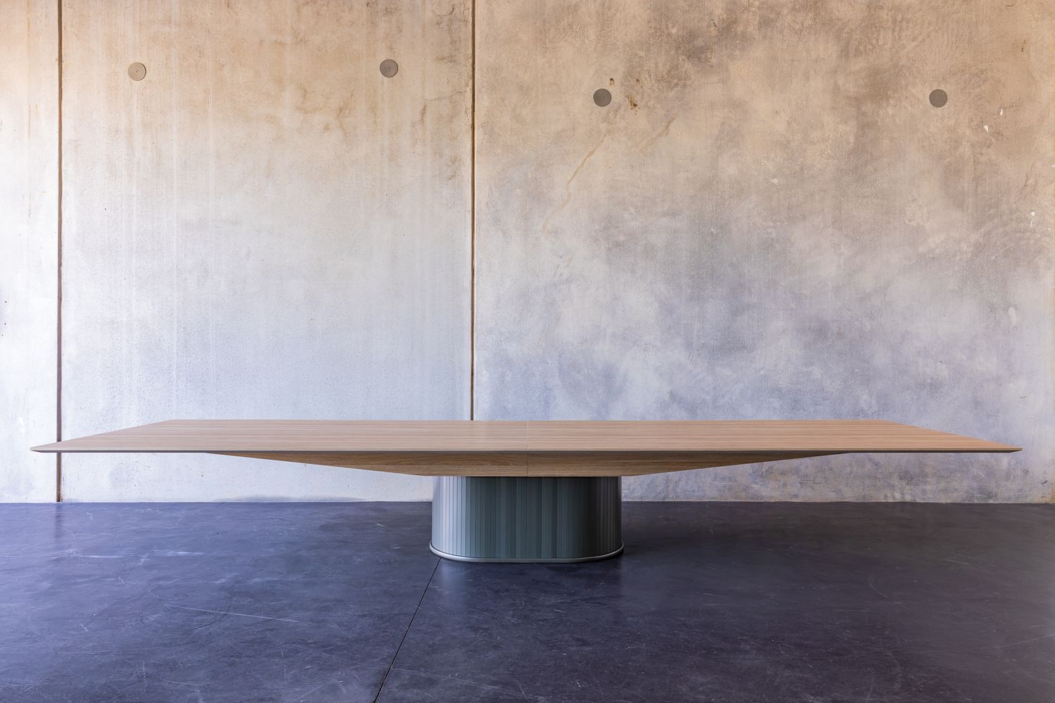 Large minimalist oak boardroom table with a sculptural tapered pedestal base in front of a concrete wall on a dark floor.