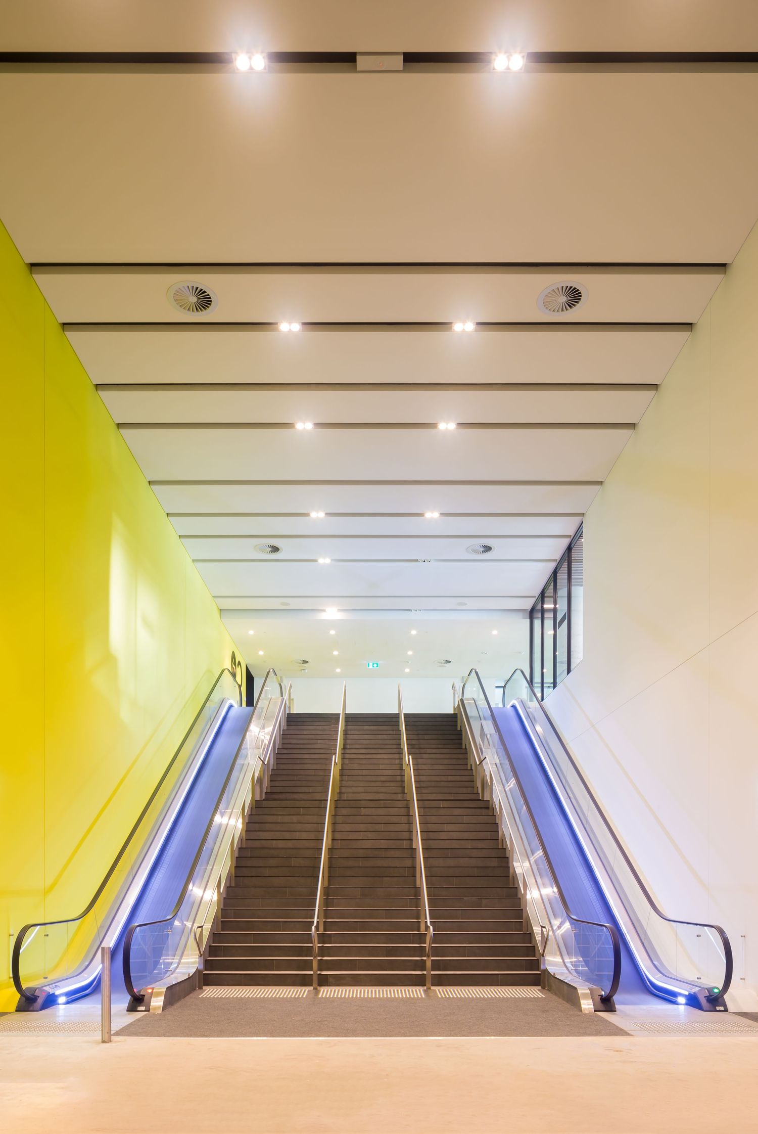Wide staircase flanked by illuminated escalators with blue lighting and a bright yellow wall in a modern convention venue.