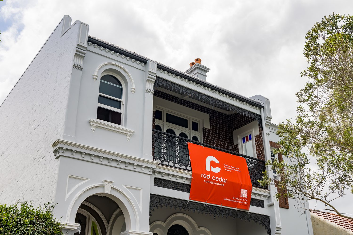 Close view of a white Victorian terrace facade with an arched window and iron balcony displaying a Red Cedar Constructions sign.