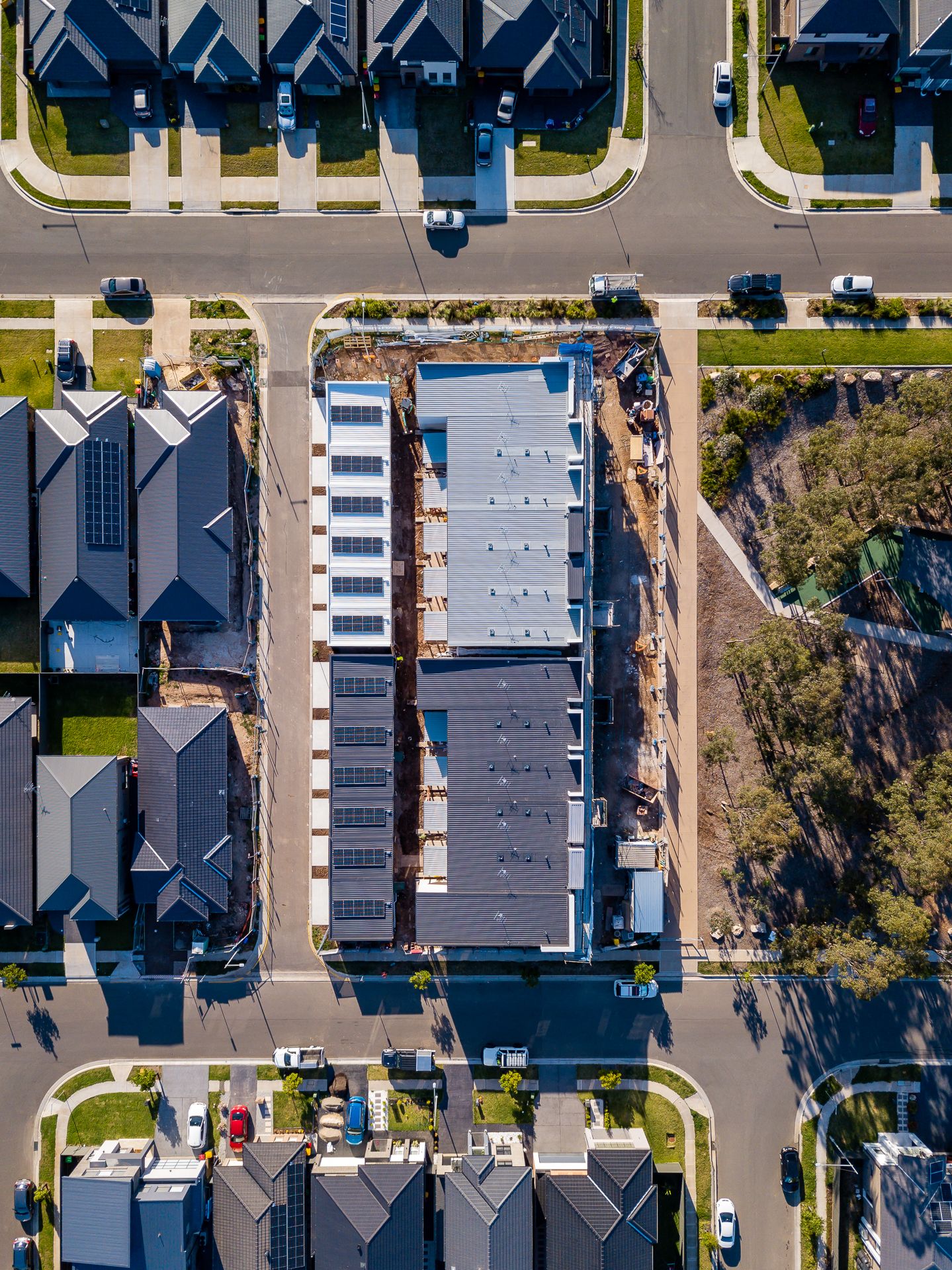 Aerial view of a row of townhouses under construction with grey rooftops among completed homes, streets and green space.