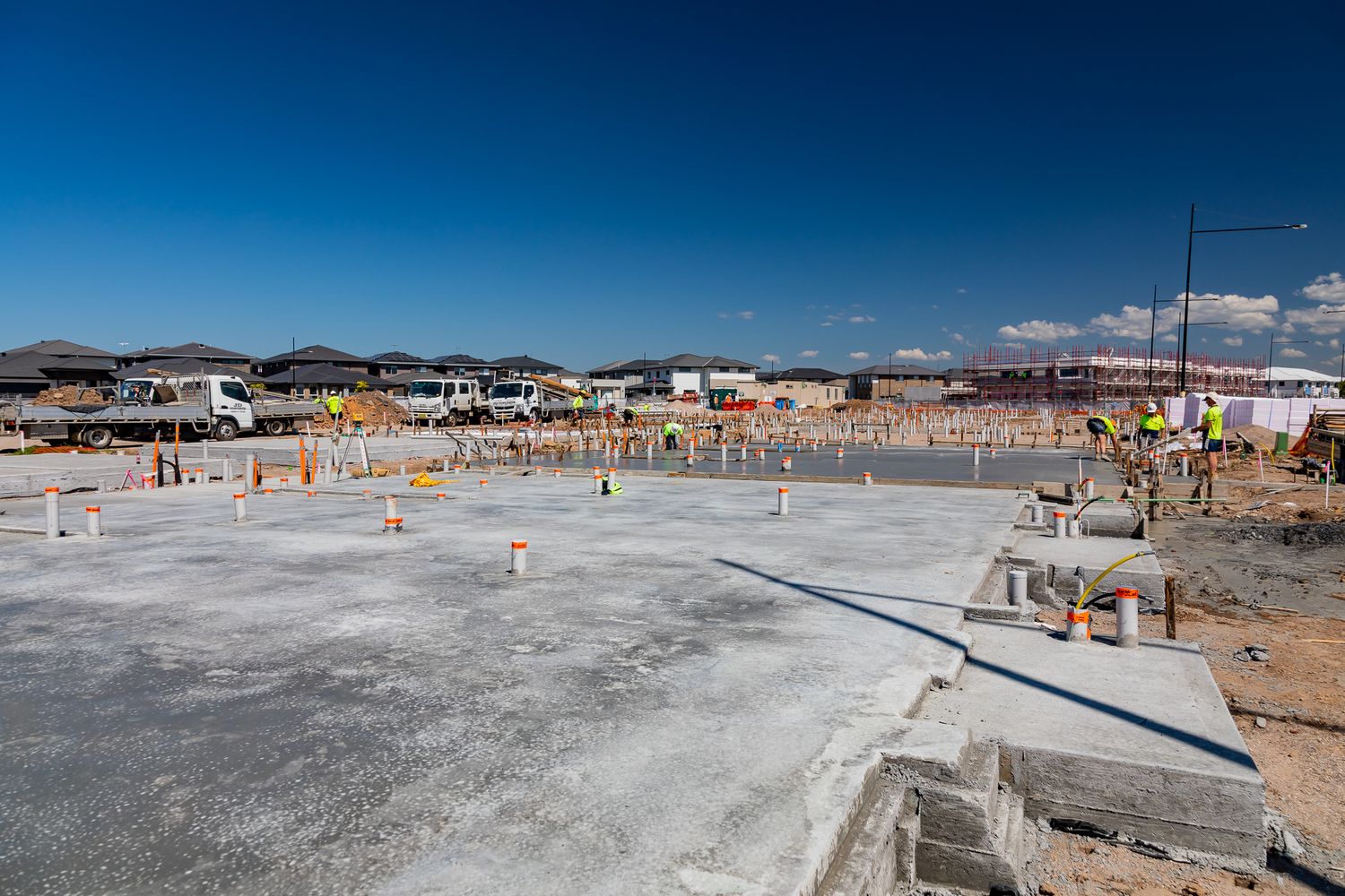 Wide view of a newly poured concrete slab with plumbing pipes and workers in the distance at a housing construction site under blue sky.