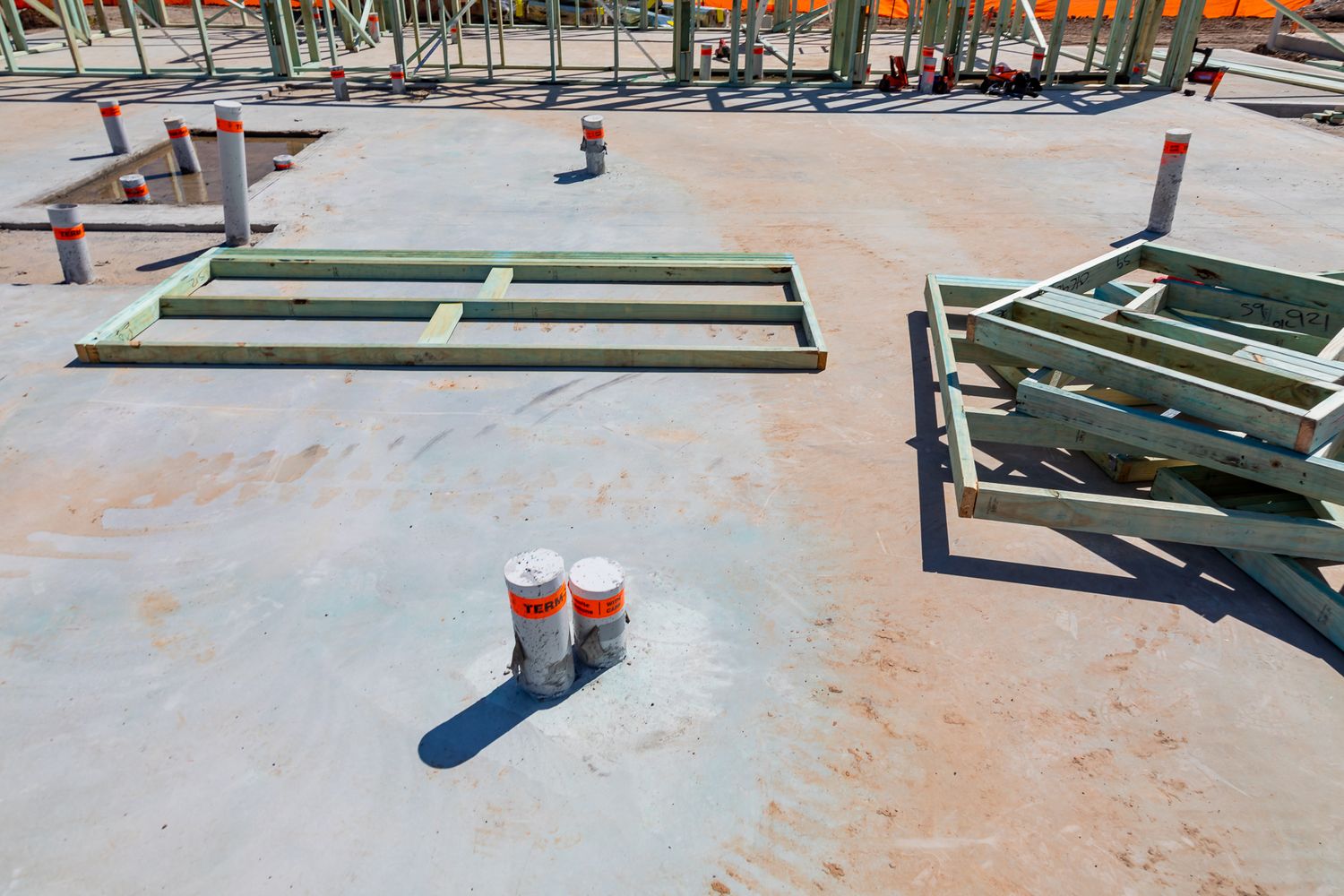 Concrete slab at a building site with timber wall frames laid flat, safety-capped rebar and steel posts awaiting framing.