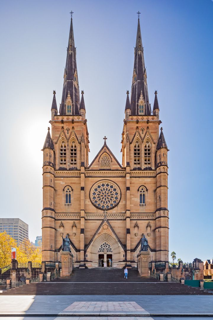Twin-spired Gothic Revival cathedral with a large rose window and sandstone facade rising above stone steps against a clear blue sky.