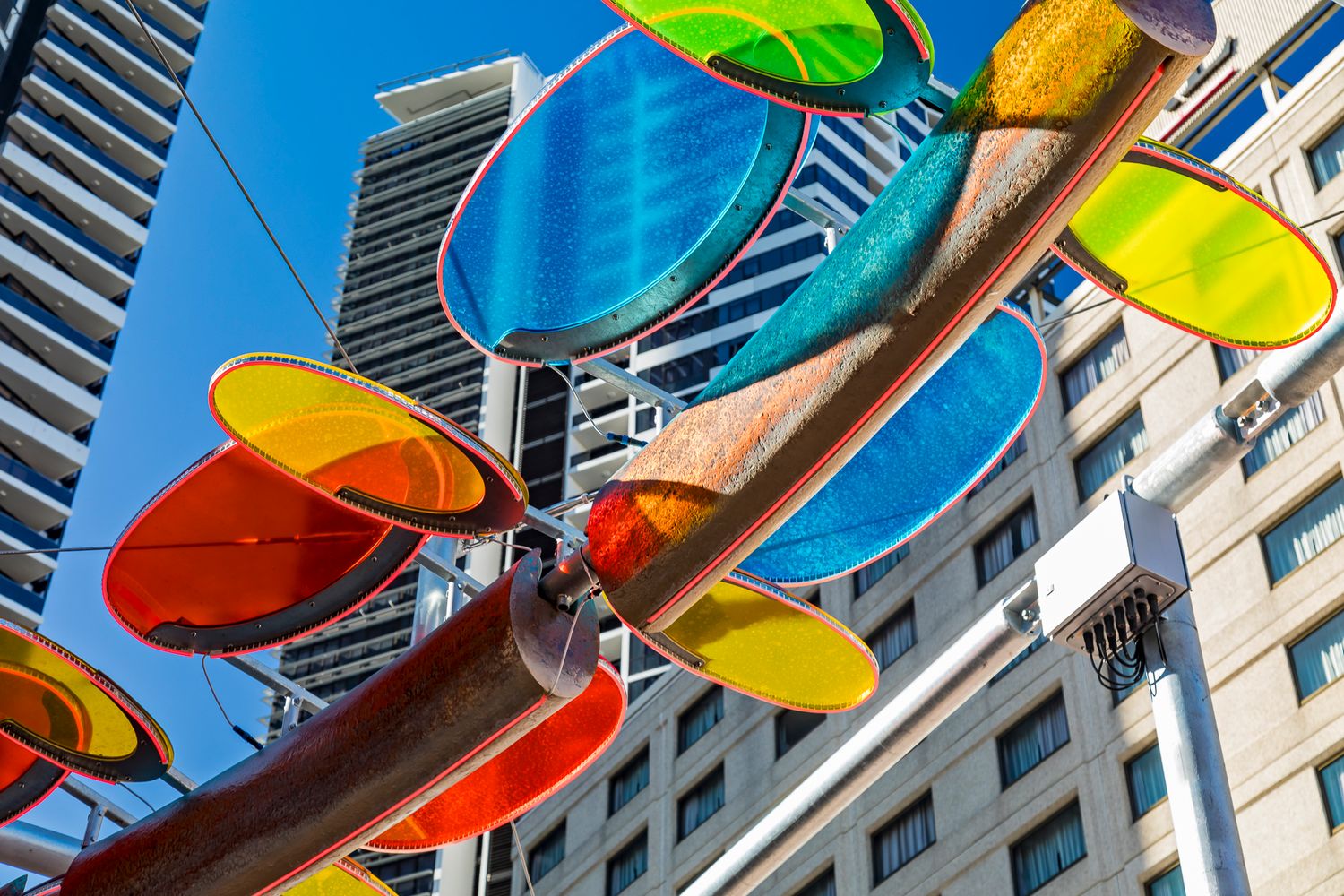 Looking up at blue, orange, red and yellow translucent discs mounted on a bronze sculpture with high-rise buildings in the background.