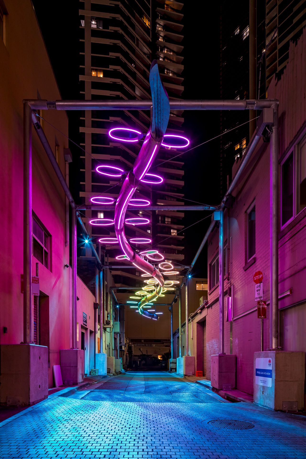 A purple neon spiral sculpture glows above a pink-lit brick alley between high-rise buildings at night.