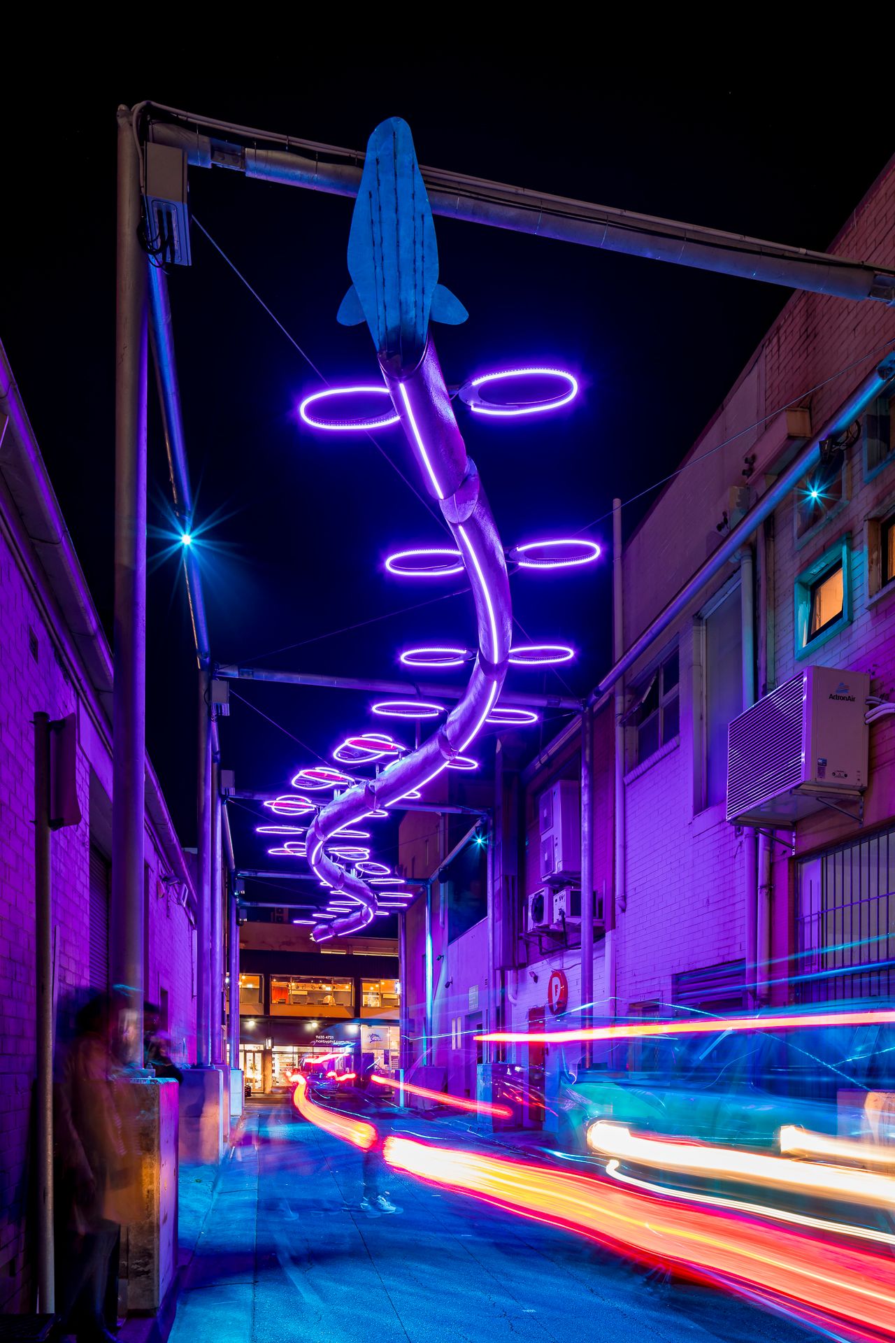Purple neon fish sculpture and glowing rings over an urban laneway at night, with red light trails from passing vehicles below.