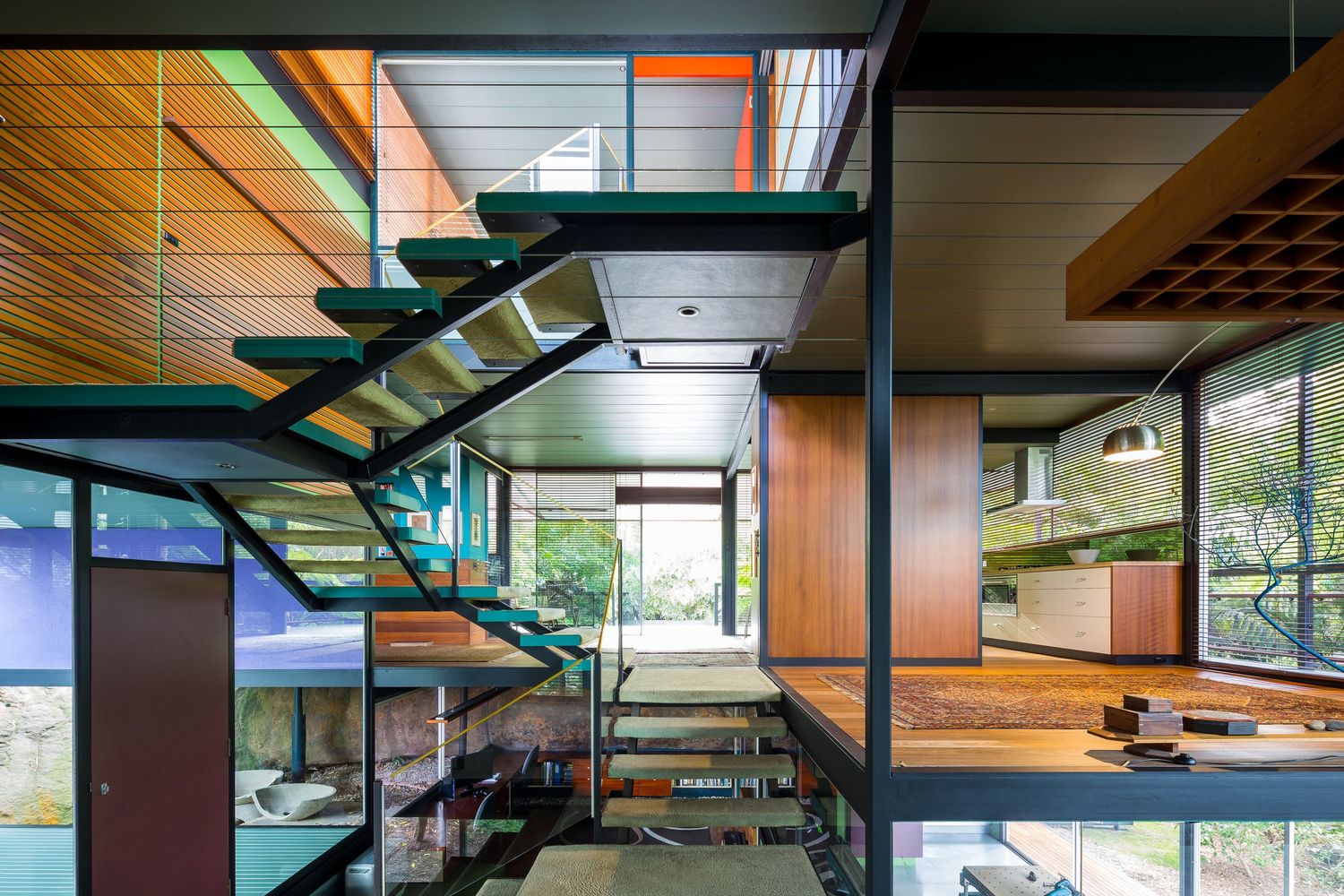 Modern timber and glass interior featuring a floating green steel staircase, wood paneling, and large windows overlooking trees.