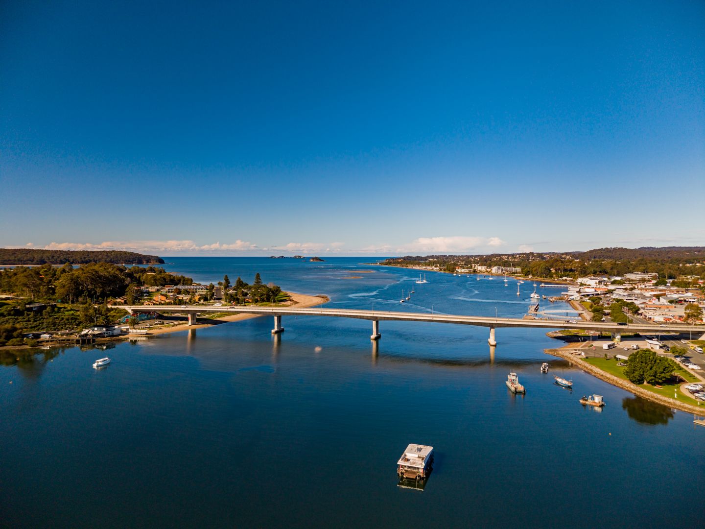 Aerial view of a long concrete bridge spanning a calm blue estuary beside a waterfront town with moored boats under a clear sky.