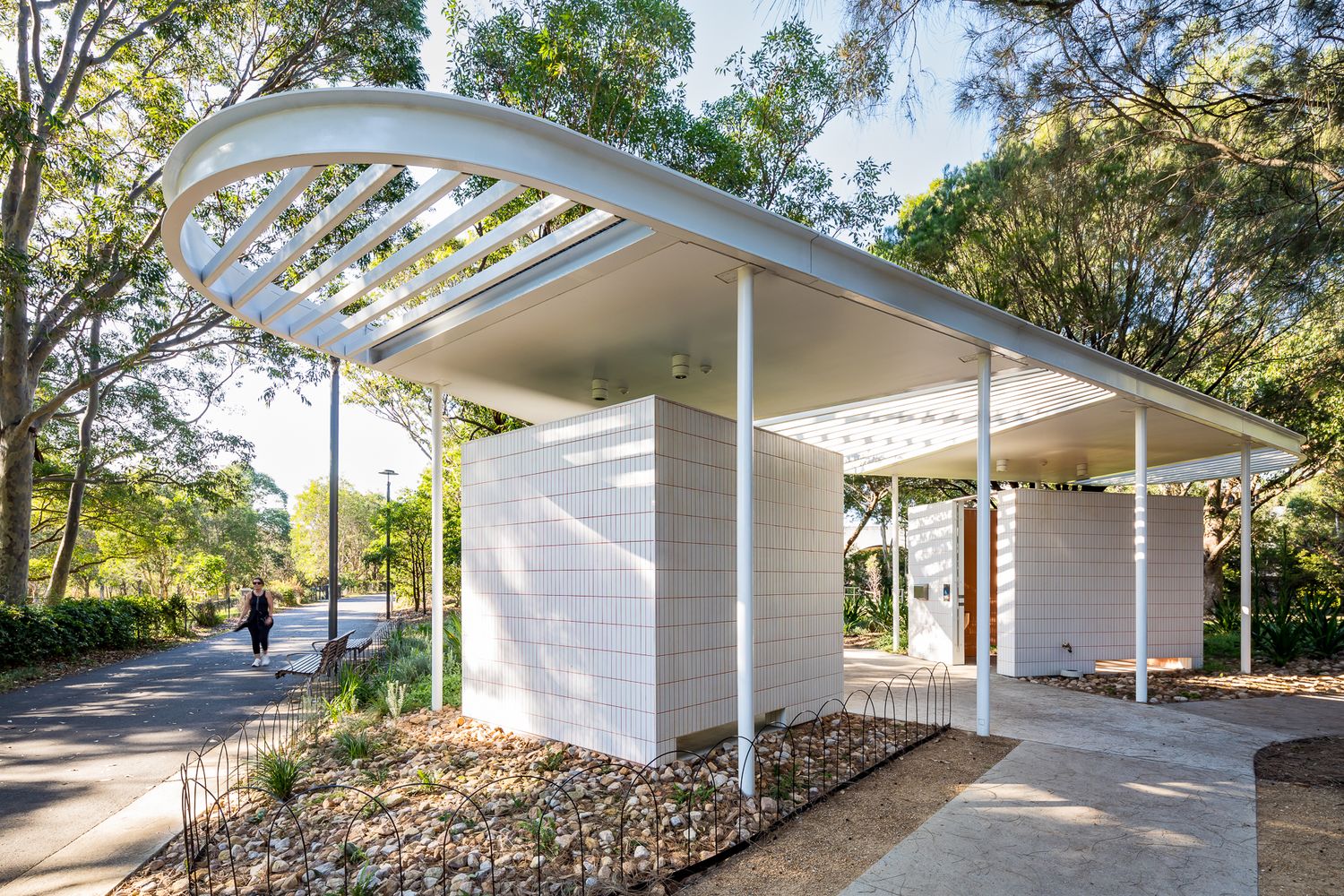 Contemporary white brick pavilion with a sweeping slatted canopy roof beside a footpath, shaded by gum trees.