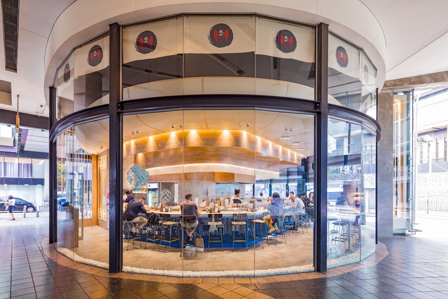 Round glass-fronted restaurant in a shopping centre with diners seated at a central counter and red circular signs above.