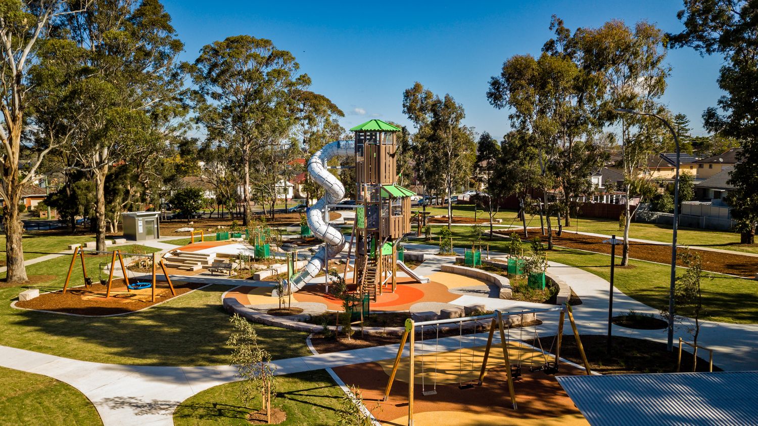 Aerial view of a modern public playground with climbing towers, slides and swings surrounded by trees, lawn and curving footpaths.