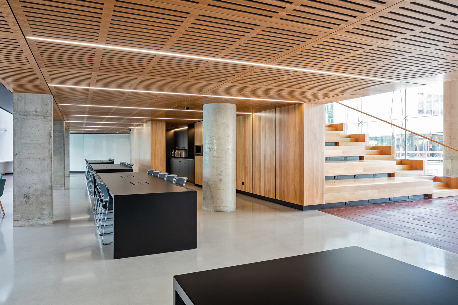 Modern open-plan interior with timber slat ceiling, concrete columns, dark communal tables and a wooden tiered seating staircase.