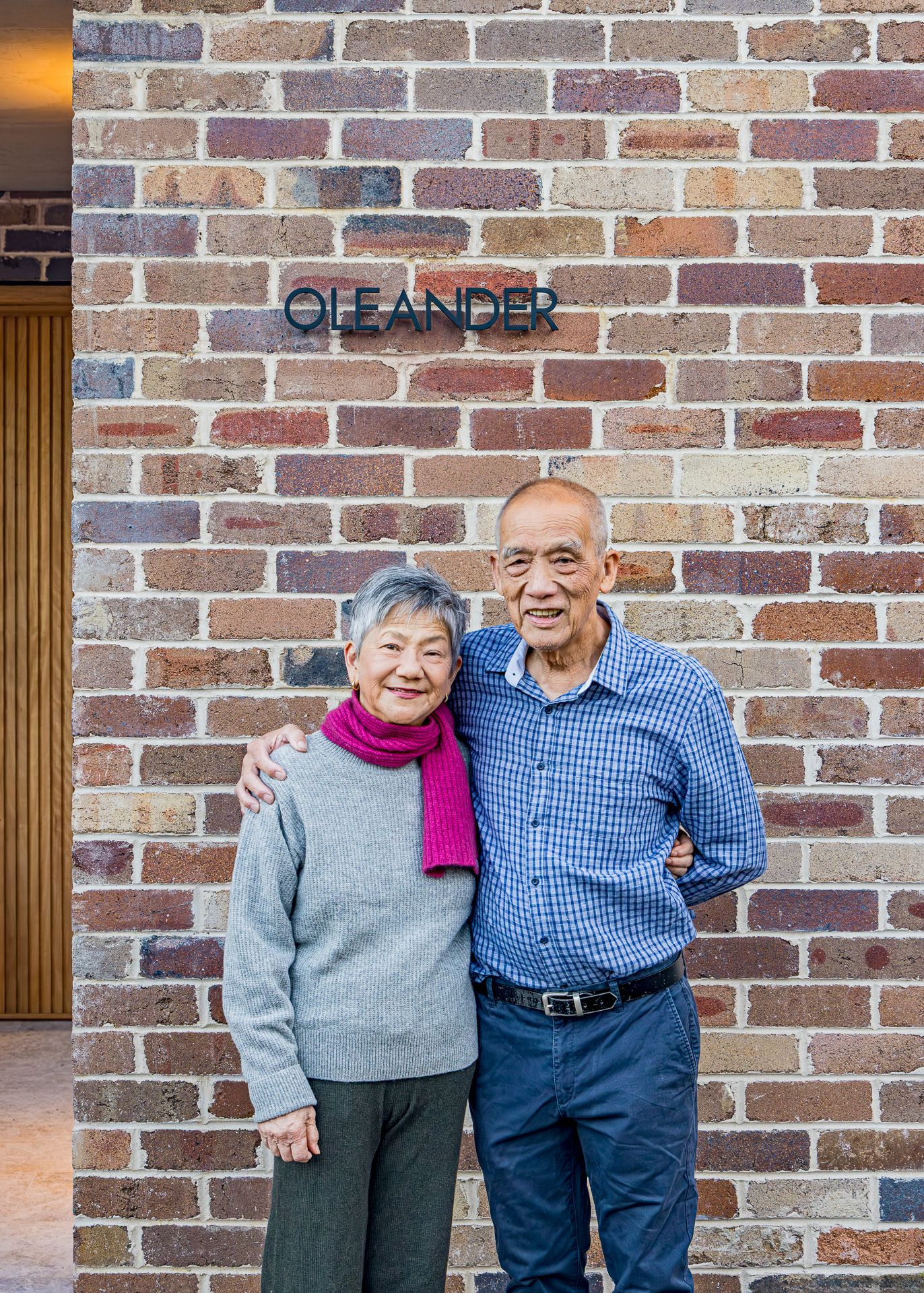 Smiling older couple embracing in front of a reclaimed brick wall with a black Oleander house sign at their new home.