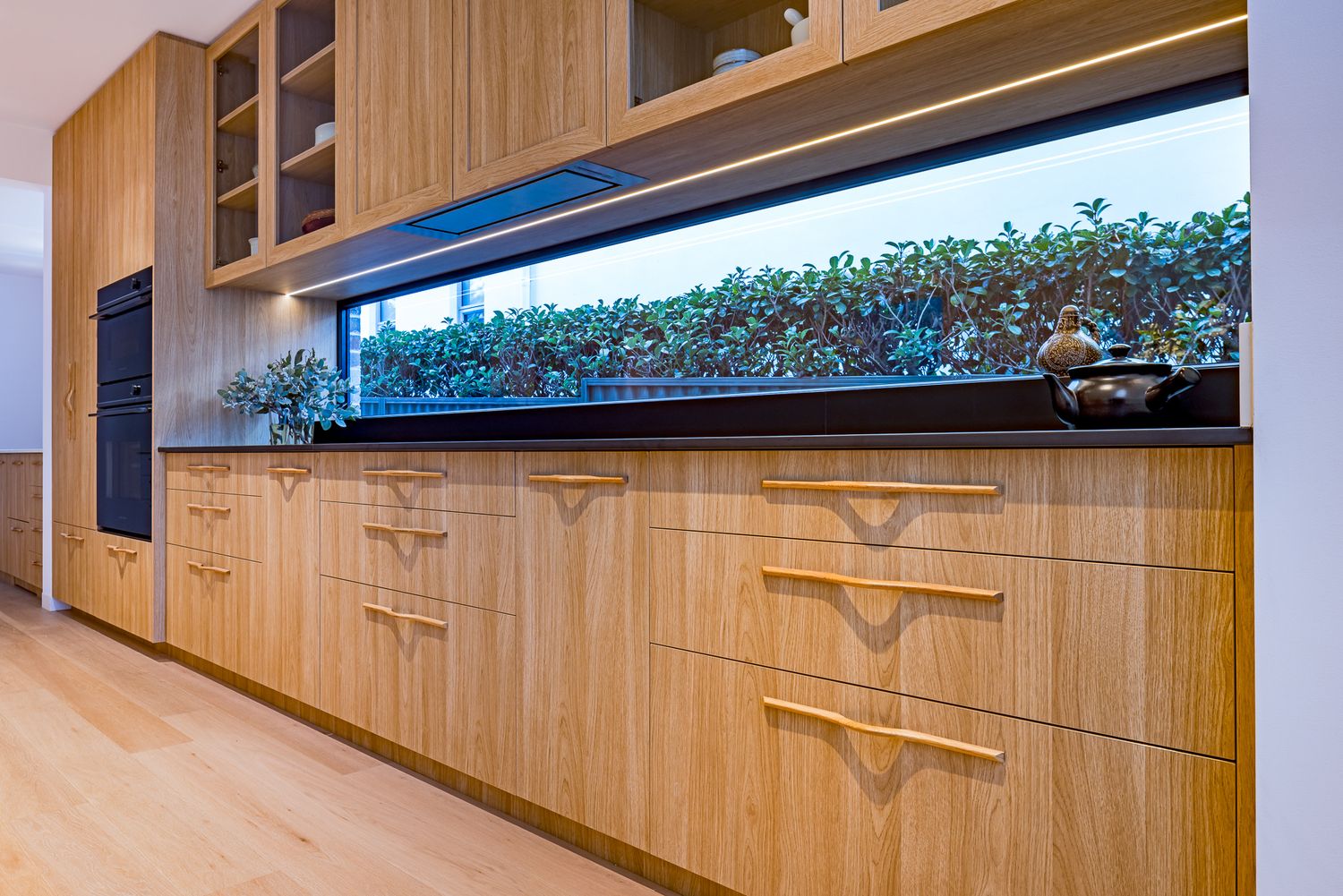 Oak kitchen cabinetry with brass handles beneath a long strip window framing a lush green hedge and styled benchtop decor.