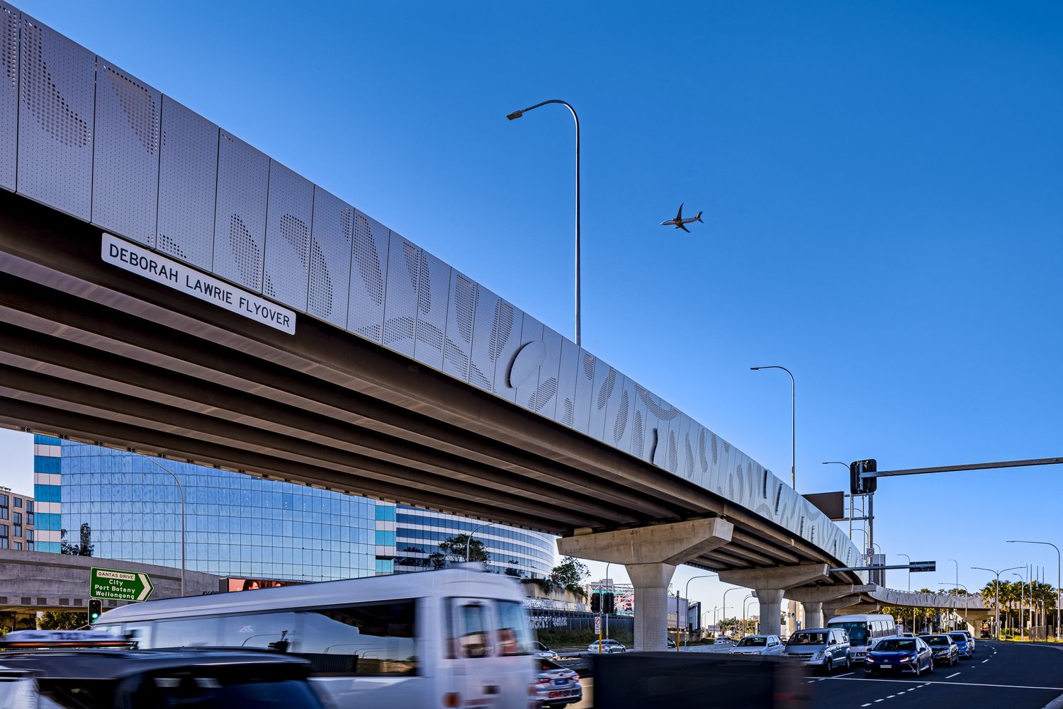 Textured concrete flyover with a street name sign above a busy road as an airplane passes overhead near modern glass buildings.
