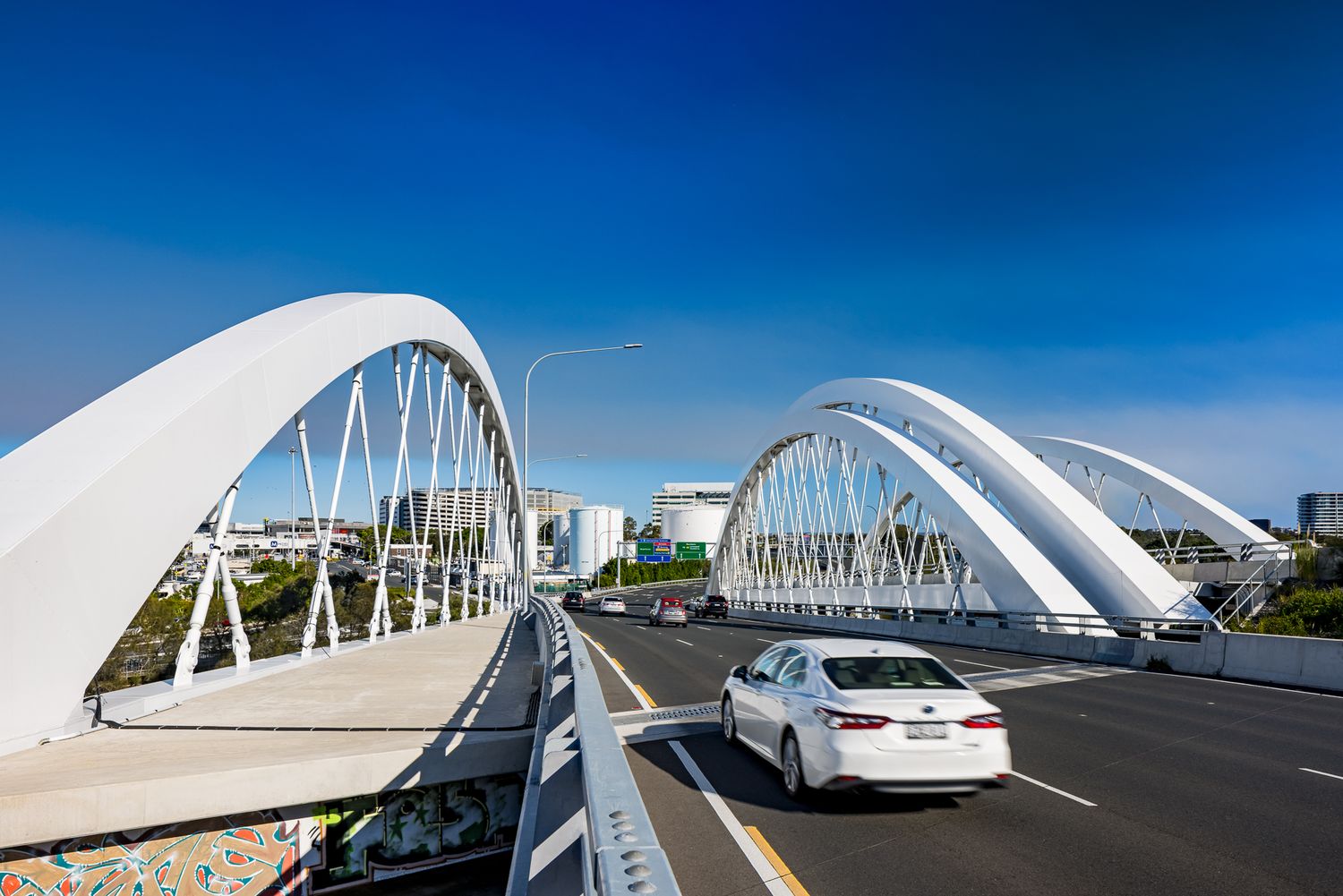 Motorway lanes passing between white steel arches of a modern bridge, with a white sedan driving under a bright blue sky.