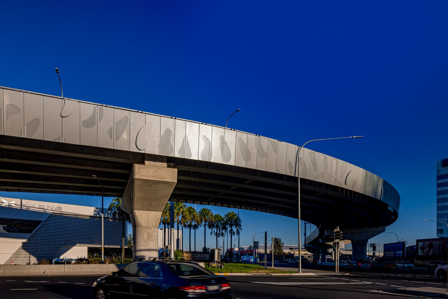 Sweeping elevated rail structure with perforated leaf-motif panels and a thick concrete pier stands above a road lined with palm trees.