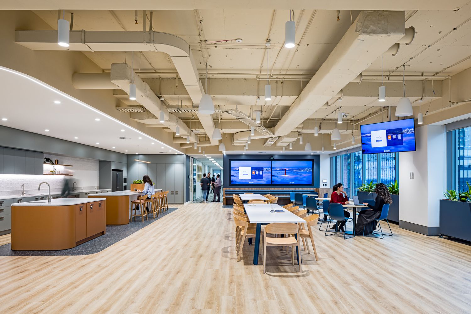 Spacious contemporary workplace cafe with light wood flooring, communal dining tables, kitchen area and city-view windows.