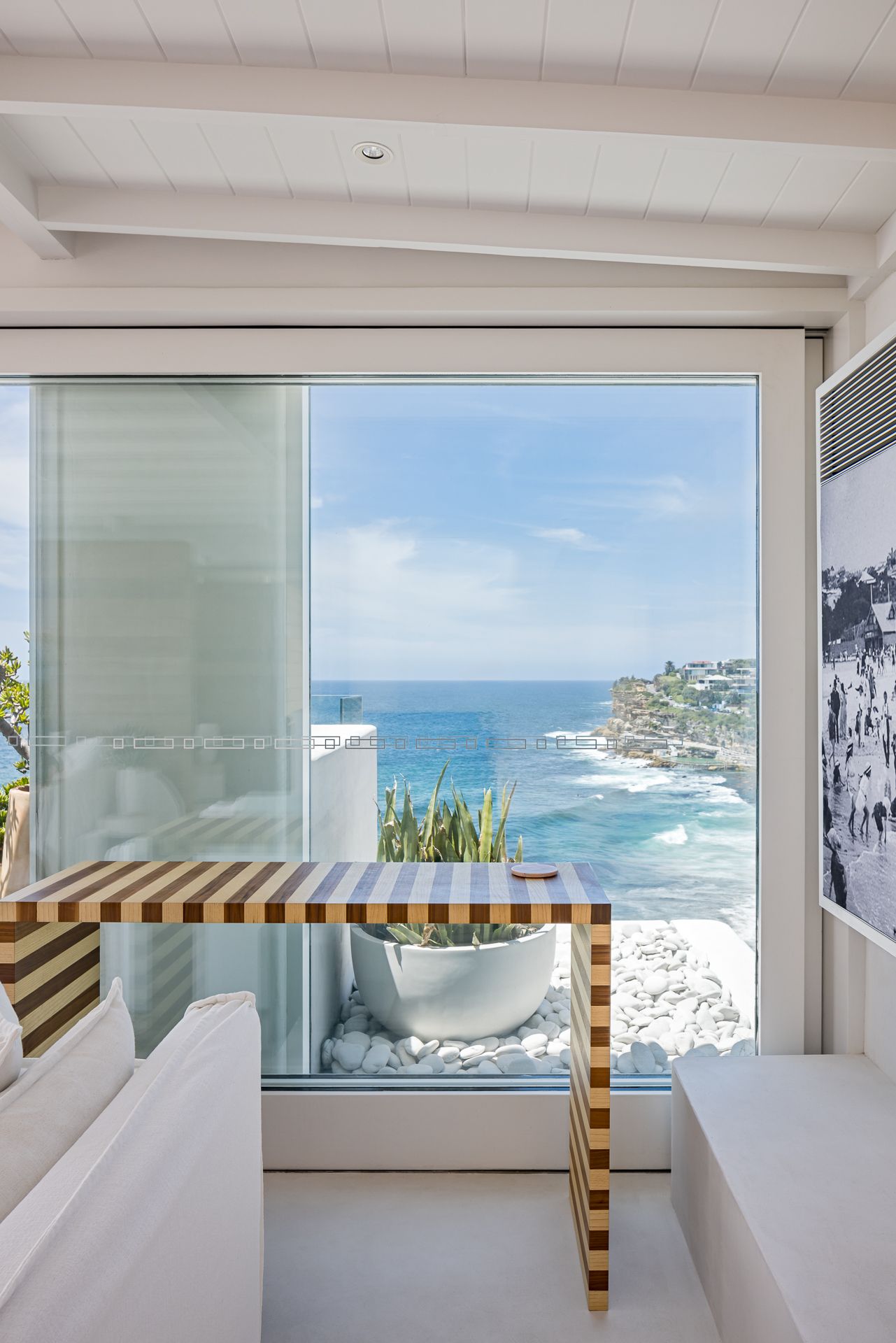 Timber striped console table beside a large window framing a sweeping ocean and coastal cliff view from a bright white interior.