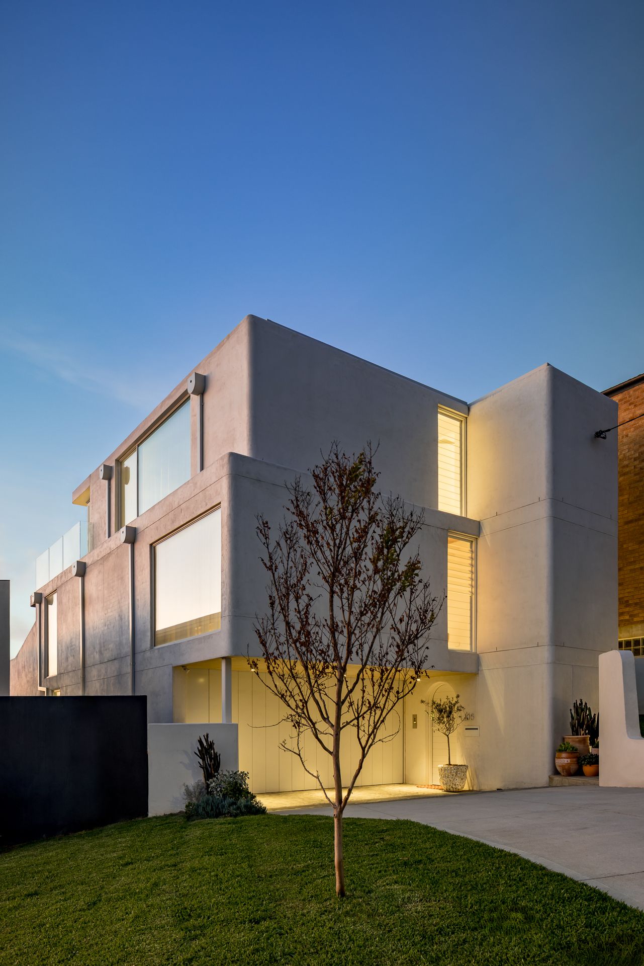 Contemporary multi-level concrete house with large windows, a young tree and a manicured lawn under a deep blue evening sky.