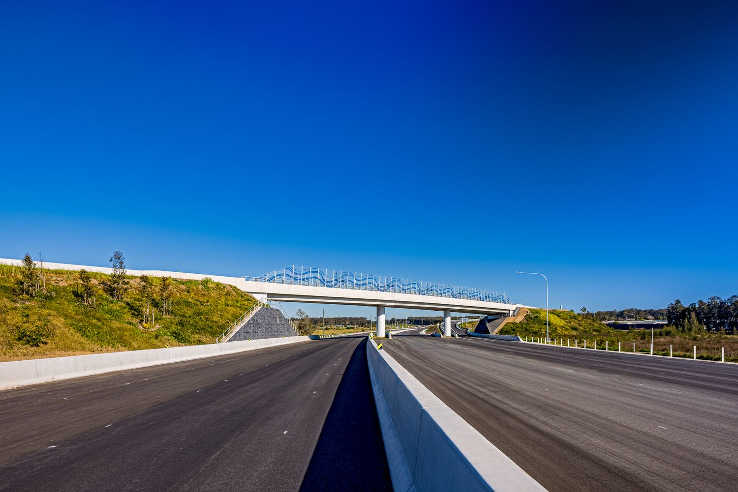 Wide view of a new multi-lane highway passing beneath a concrete overpass, with grassy embankments and clear blue sky.