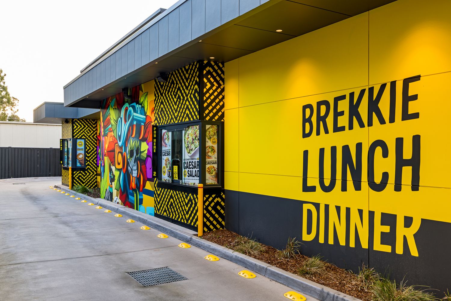 Yellow restaurant wall with bold Brekkie Lunch Dinner lettering beside a colorful graffiti mural and menu boards along the drive-thru.