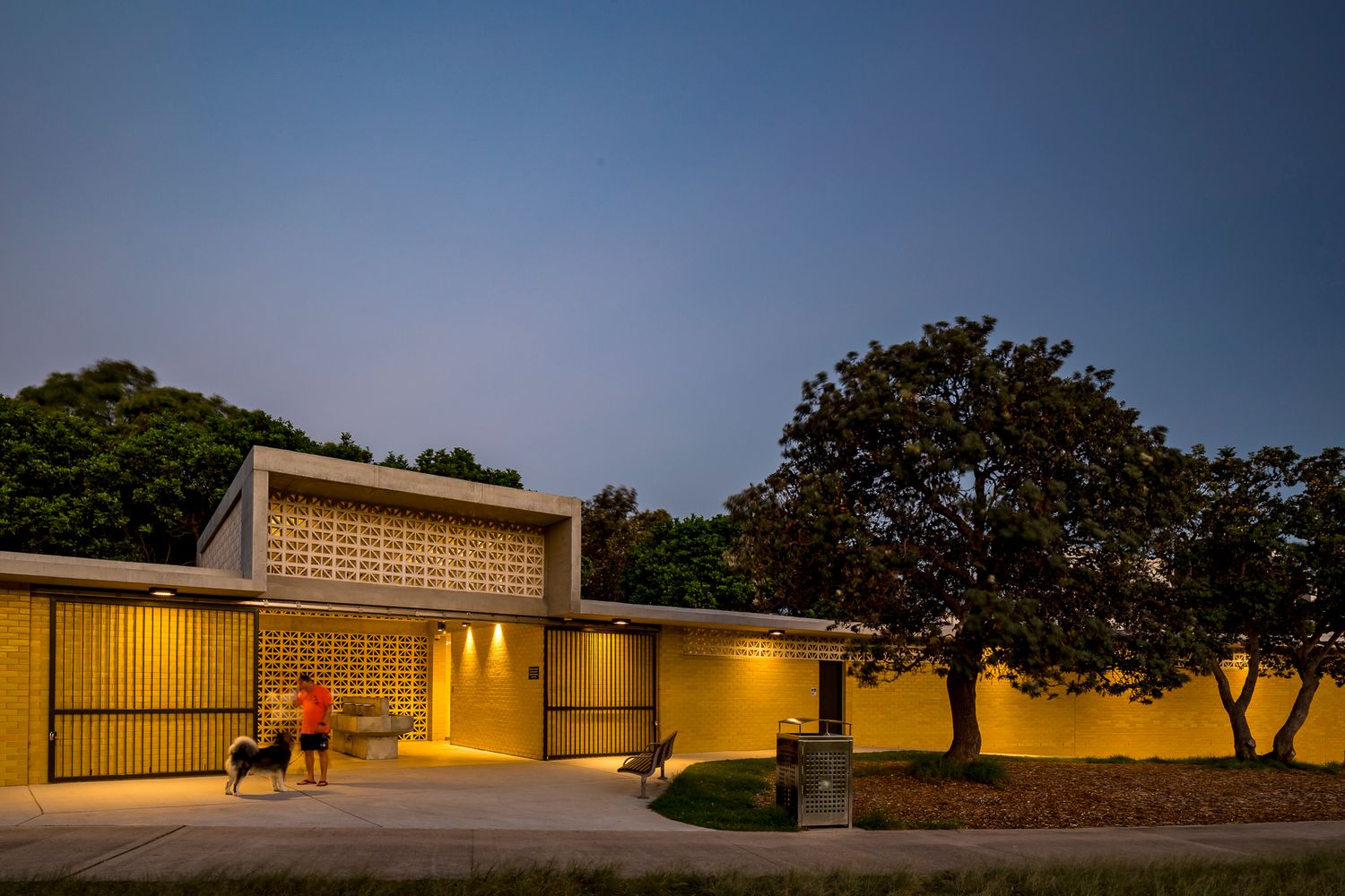 At dusk a person and a dog stand outside a softly lit yellow brick amenities building with breeze block screens and mature trees nearby.