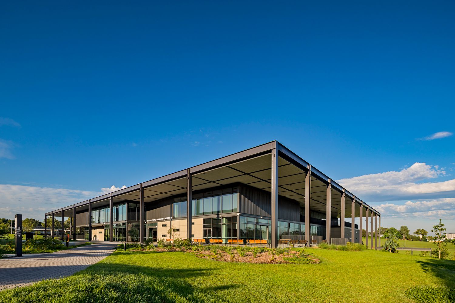 Modern TAFE campus building with a wide steel canopy, glass facade and green grounds beneath a blue sky with scattered clouds.